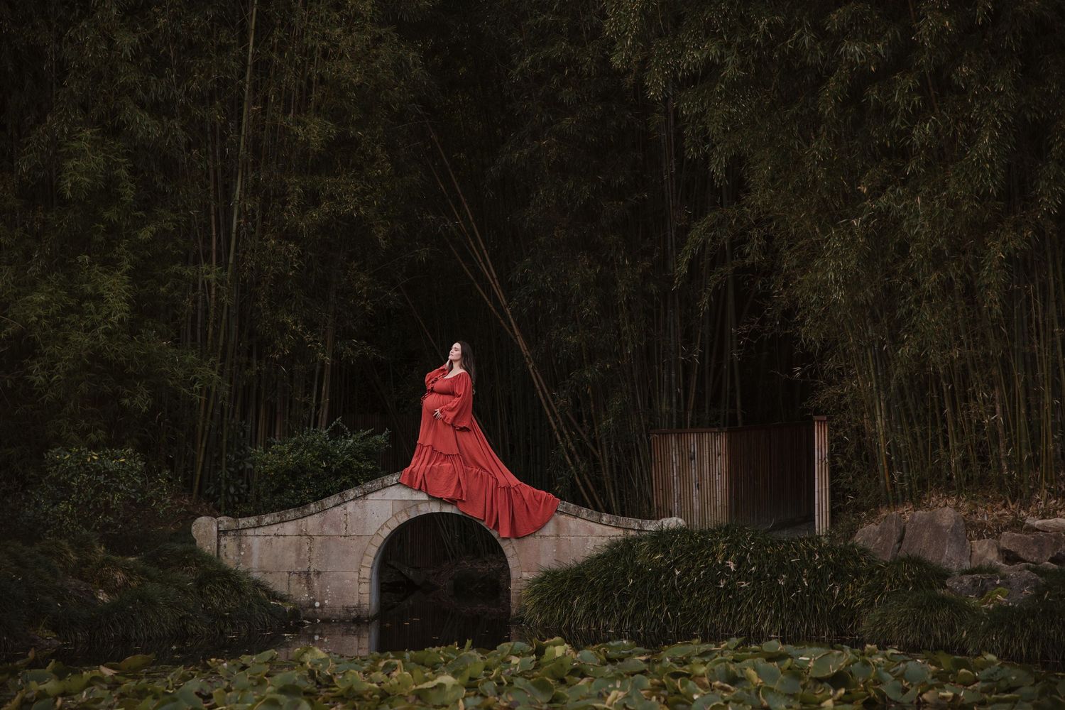 Woman in red dress stands on stone bridge surrounded by bamboo forest and lily pond.