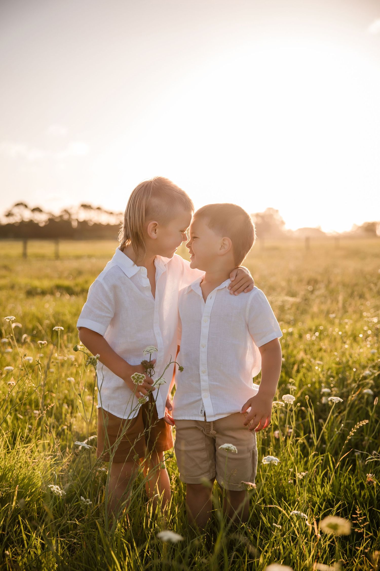 Two young boys in white shirts share a tender moment in a sunlit meadow filled with wildflowers.