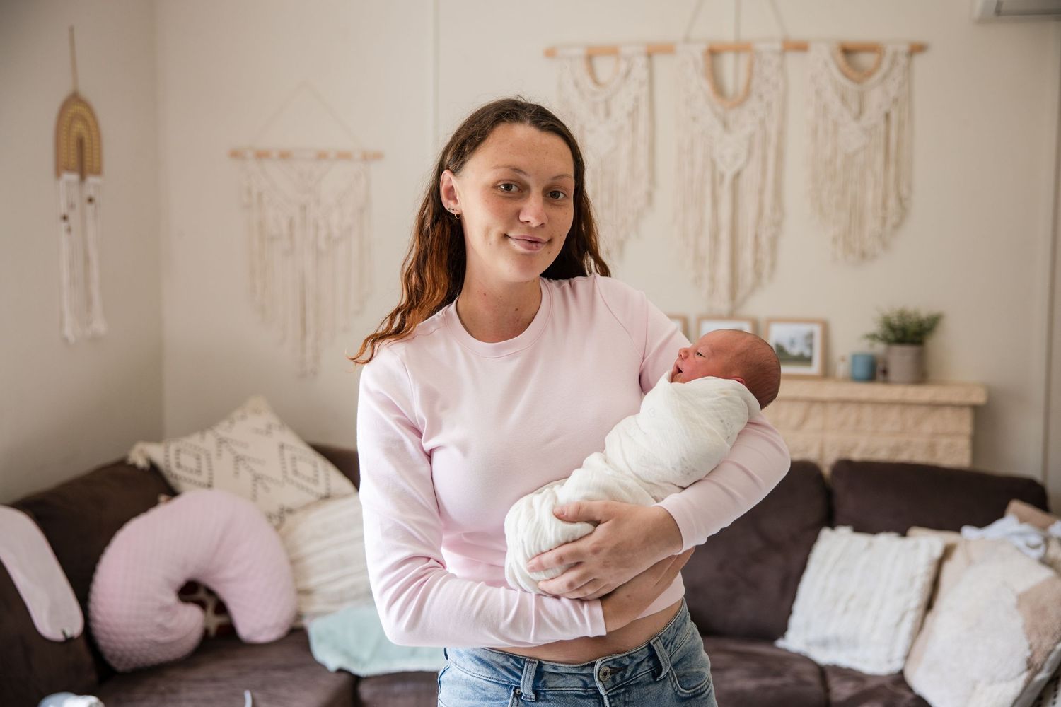Young mother in pink top holds swaddled newborn baby in cozy living room with macrame wall decor.