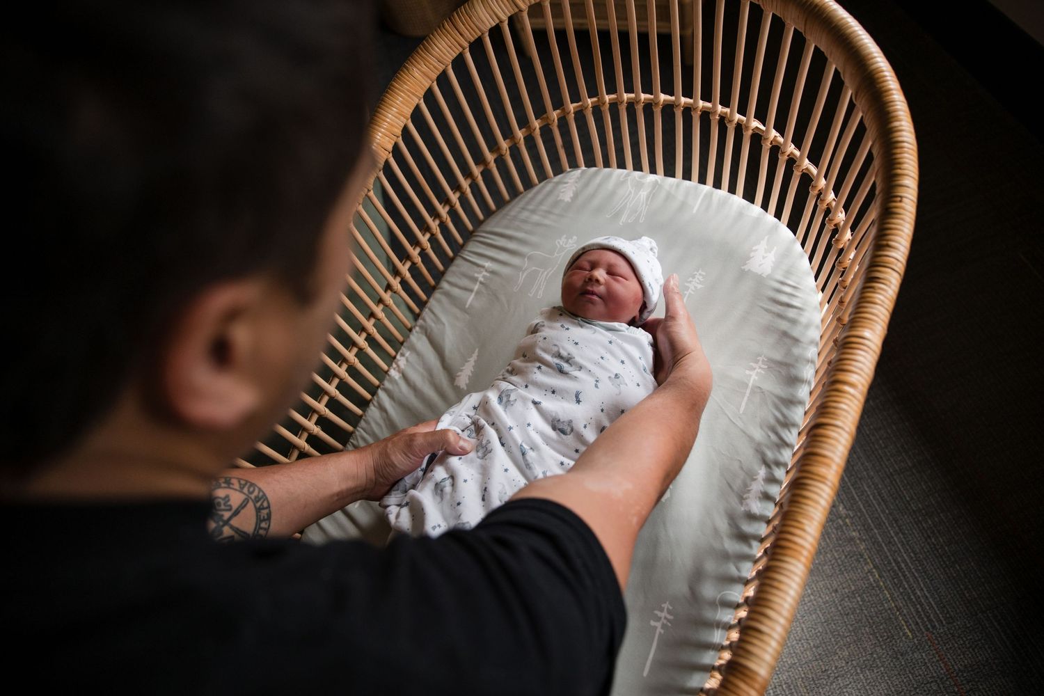 Parent gently touching newborn baby swaddled in white blanket lying in a wicker bassinet.