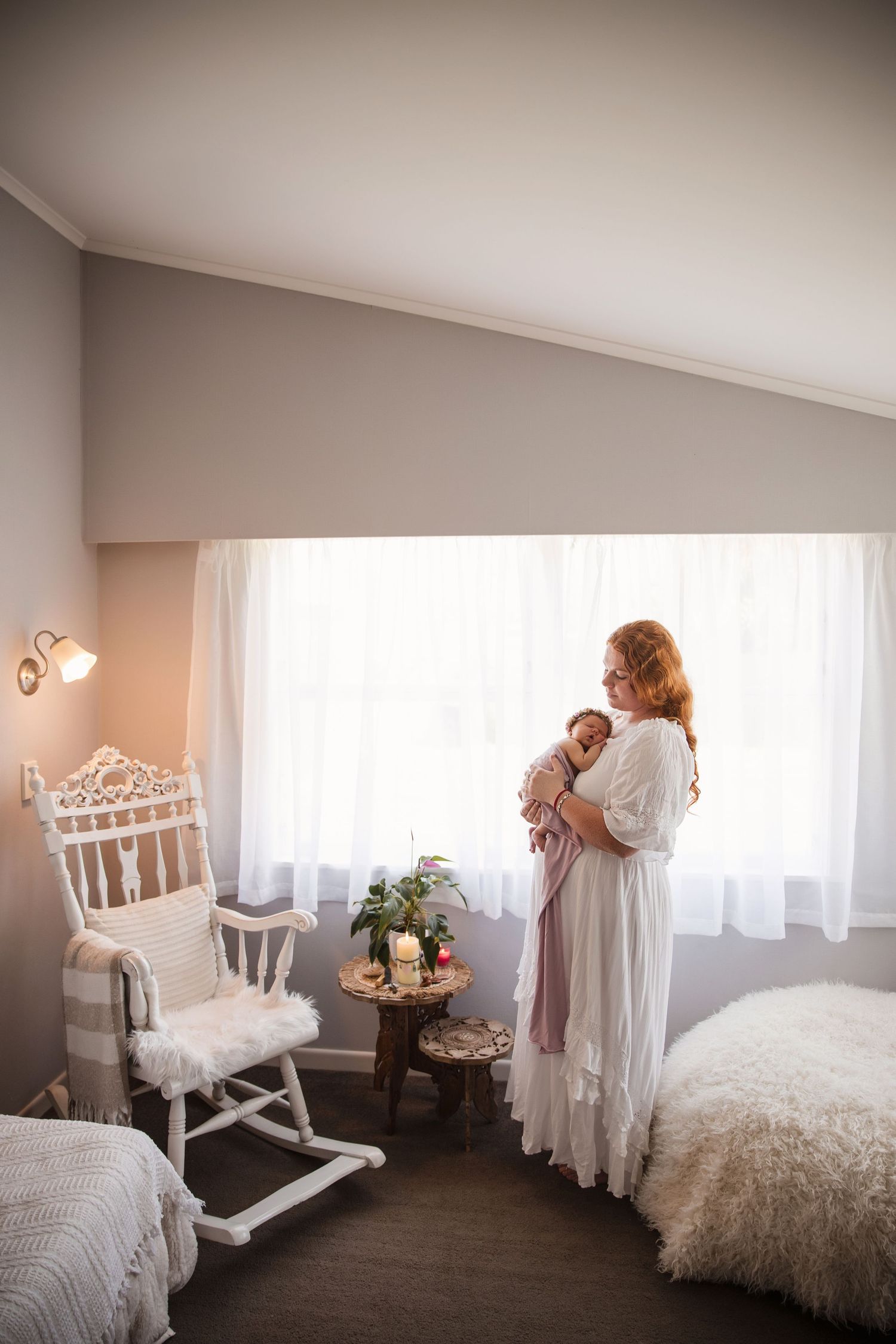 A red-haired mother in a white dress holds her newborn baby by a bright window in a serene nursery with a rocking chair.