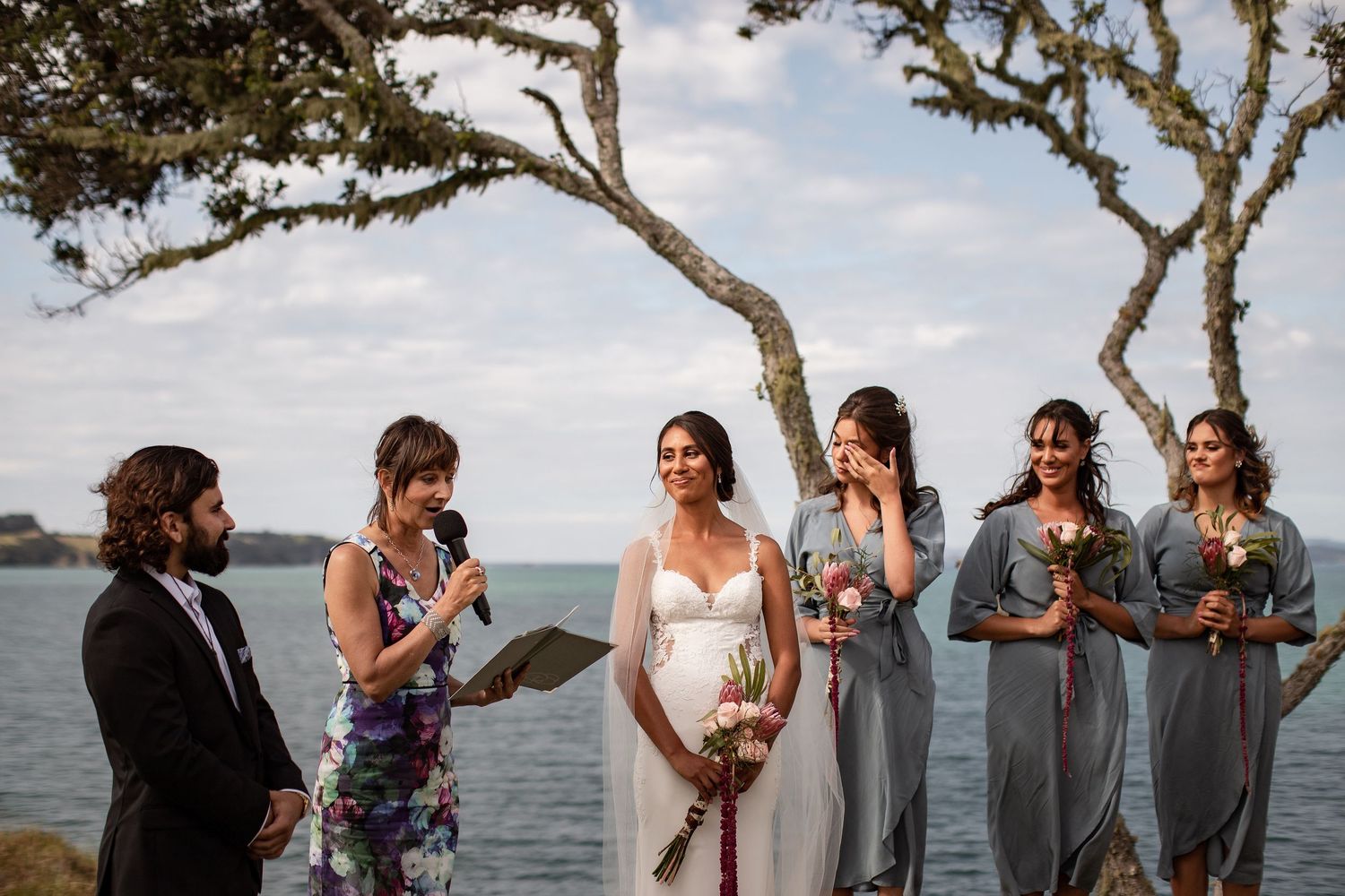 Wedding party gathered beneath twisted trees at seaside ceremony with blue water in background.