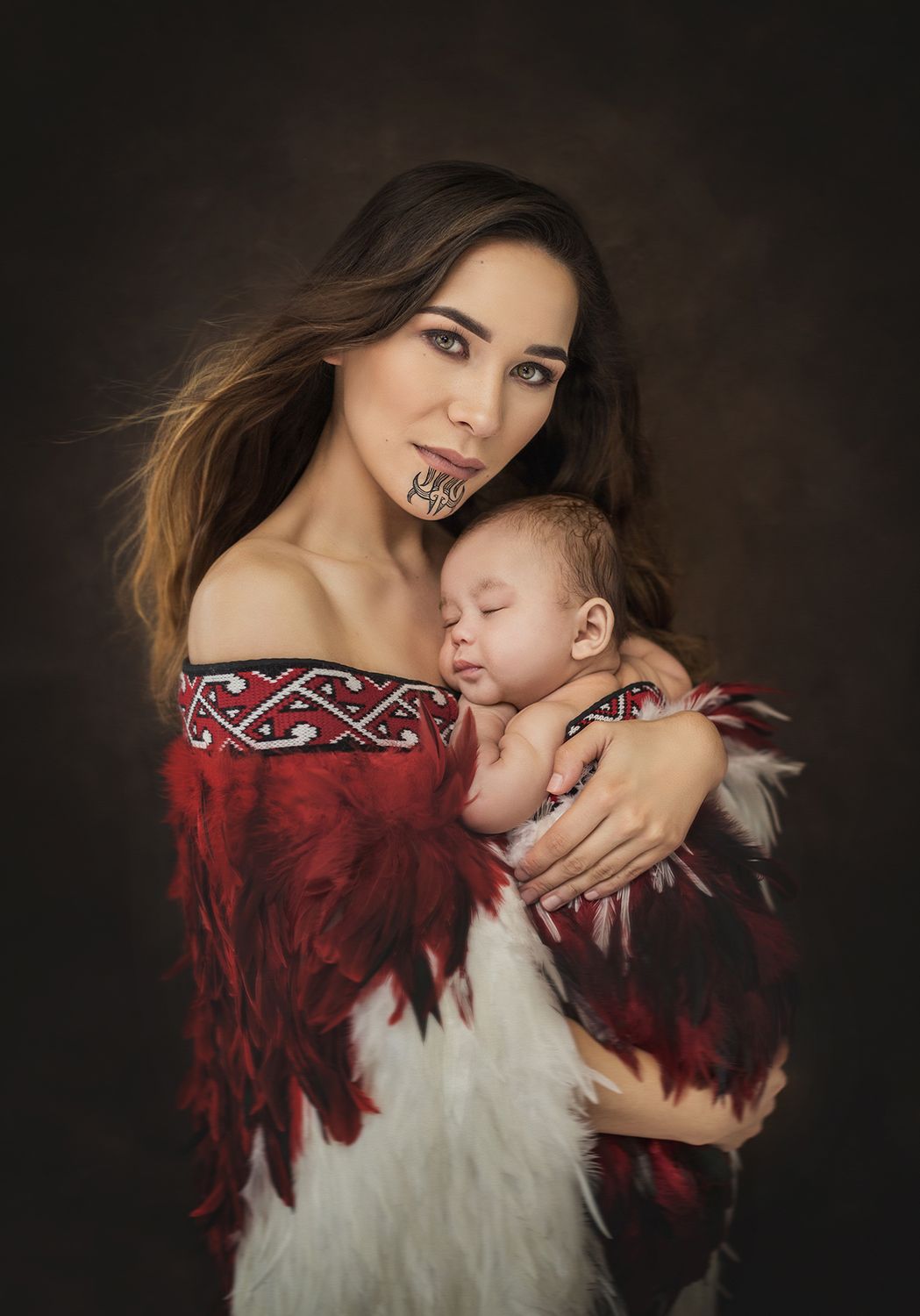 A mother in a red and white traditional dress with embroidered trim holds a sleeping baby against a dark backdrop.