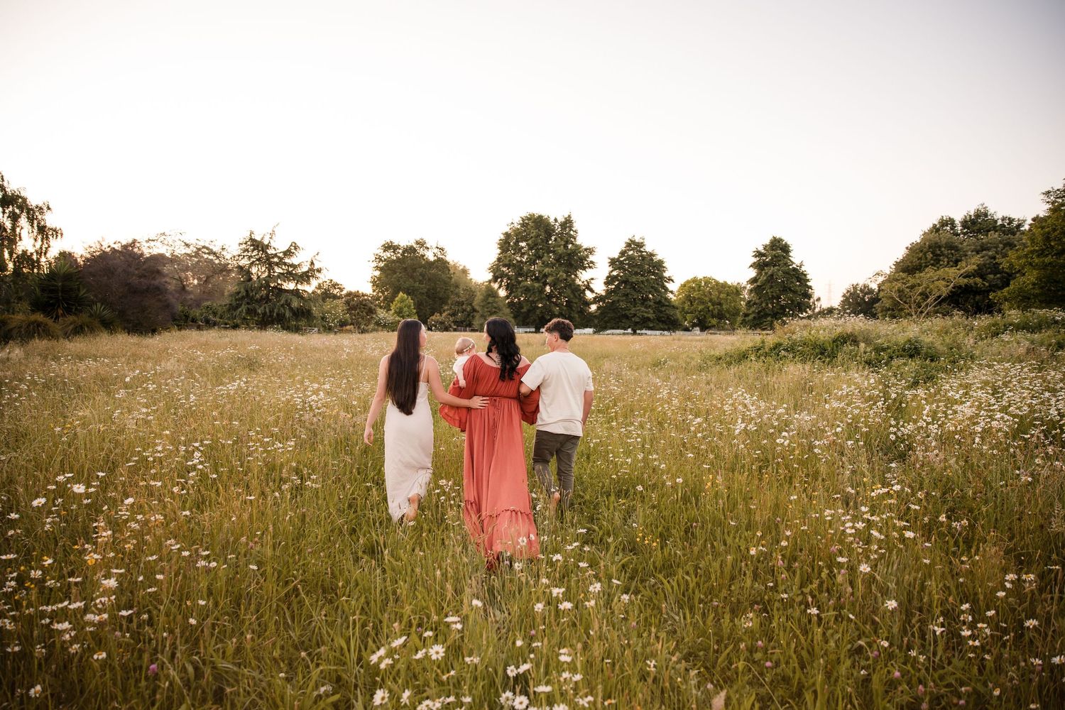 Three people walk through a sunlit wildflower meadow with trees in the background during golden hour.