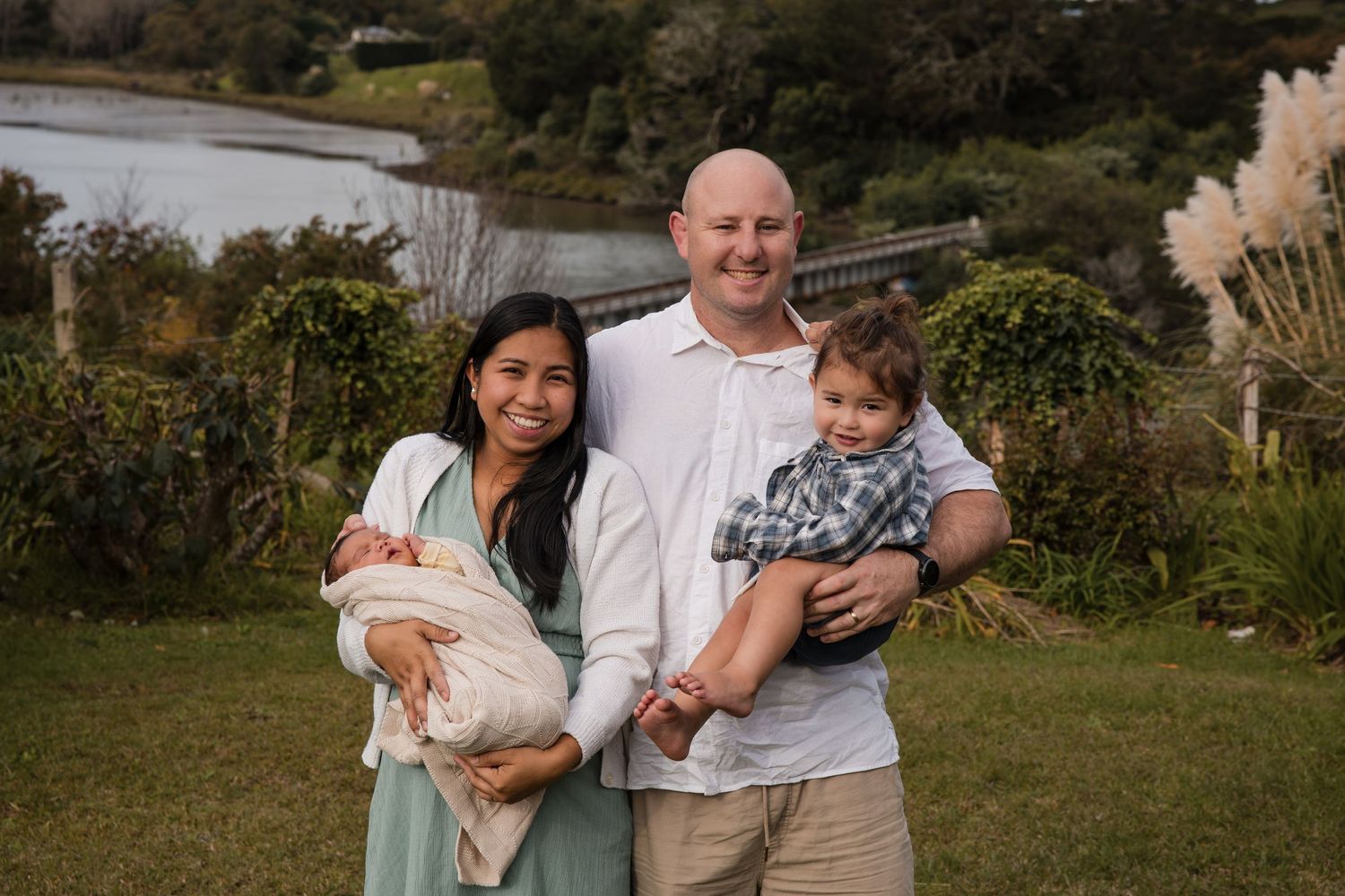 Happy family of four posing outdoors near a lake, parents holding two young children, smiling in a lush green setting.