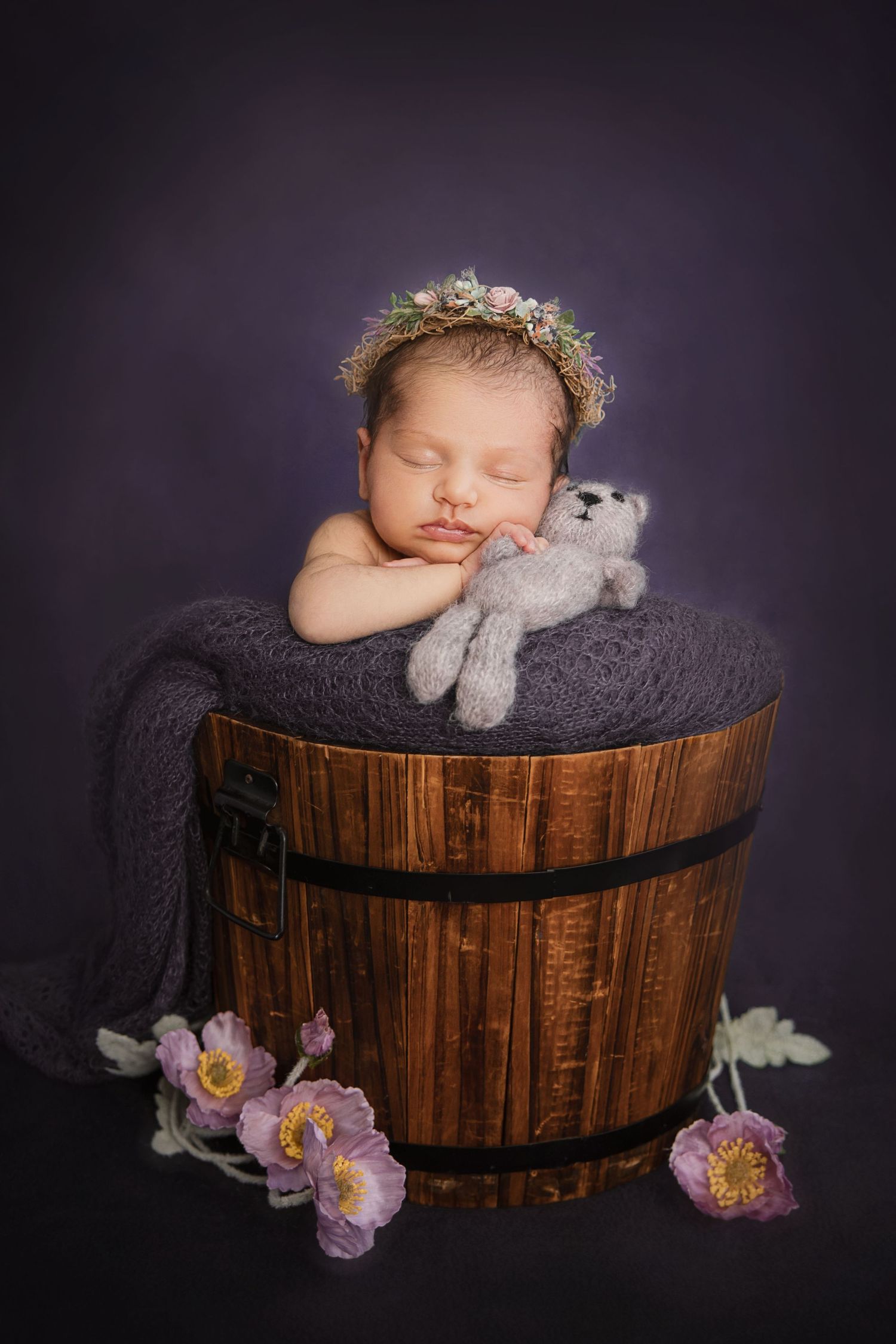 Newborn baby sleeping in rustic wooden bucket with floral crown and knit teddy bear on purple background.