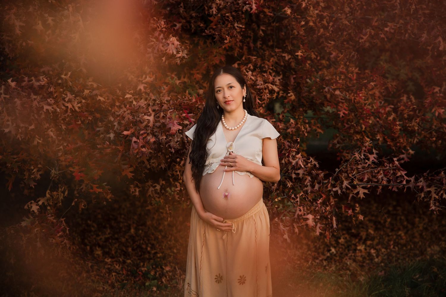 Pregnant woman in white top and beige skirt cradling baby bump outdoors among red autumn foliage.