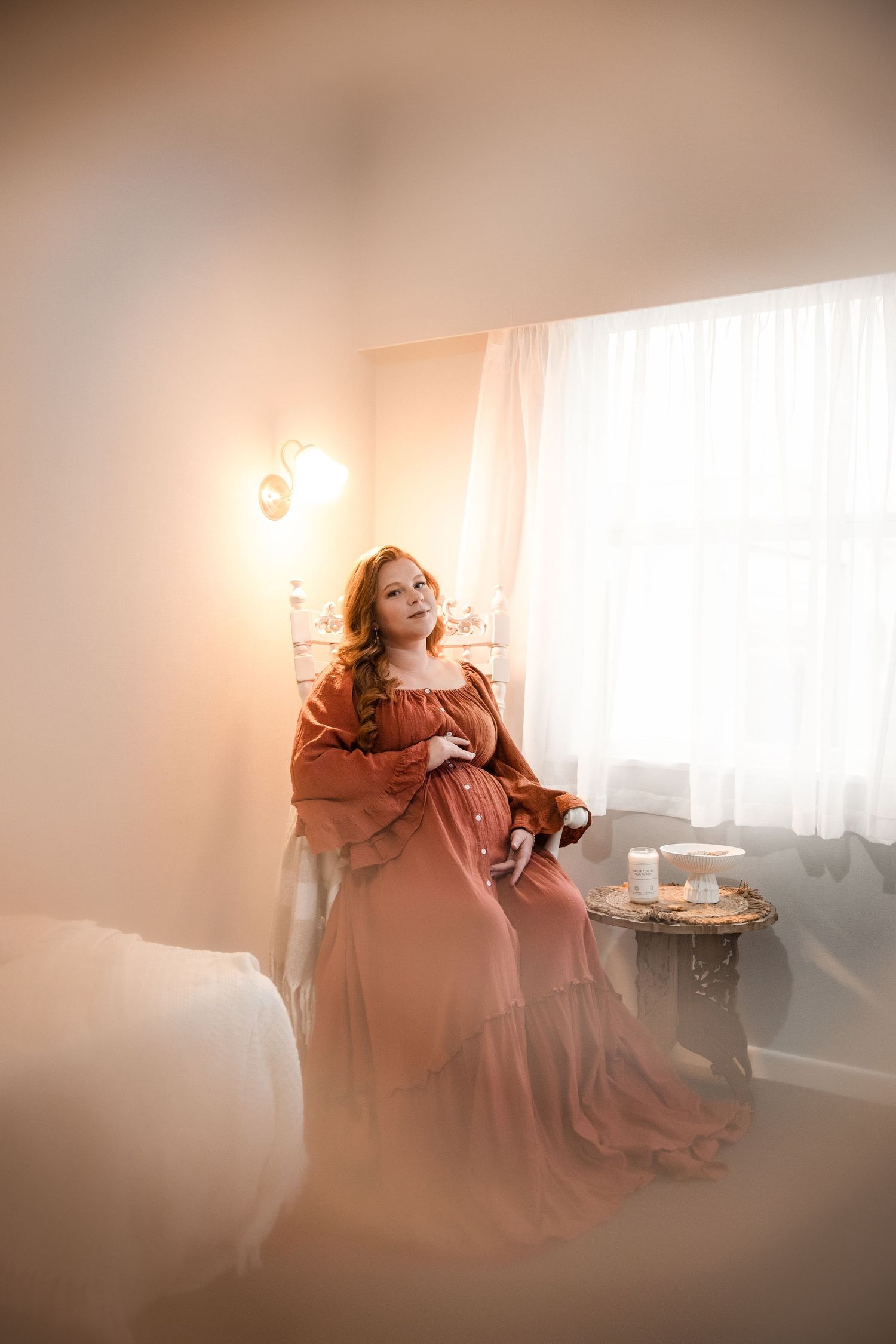A pregnant woman in a flowing rust-colored gown sits gracefully by a sunlit window in a softly lit, dreamy room.