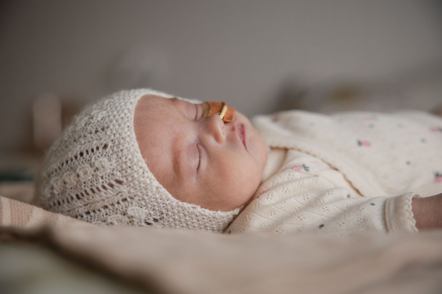 Peaceful newborn baby sleeping in white knit bonnet and floral outfit on soft bedding.