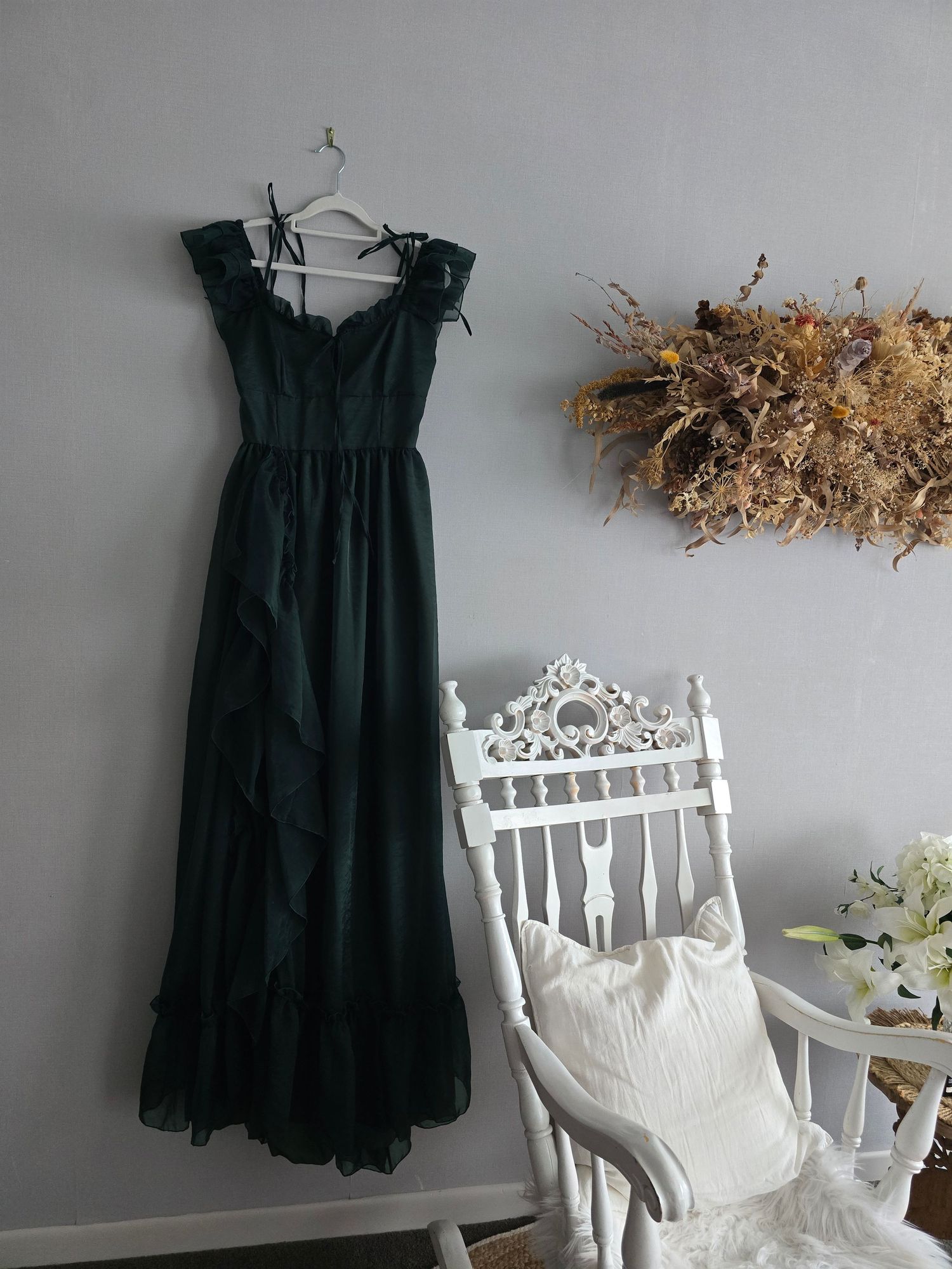 Dark green chiffon maxi dress with ruffles hanging on wall beside white ornate chair and dried floral arrangement.