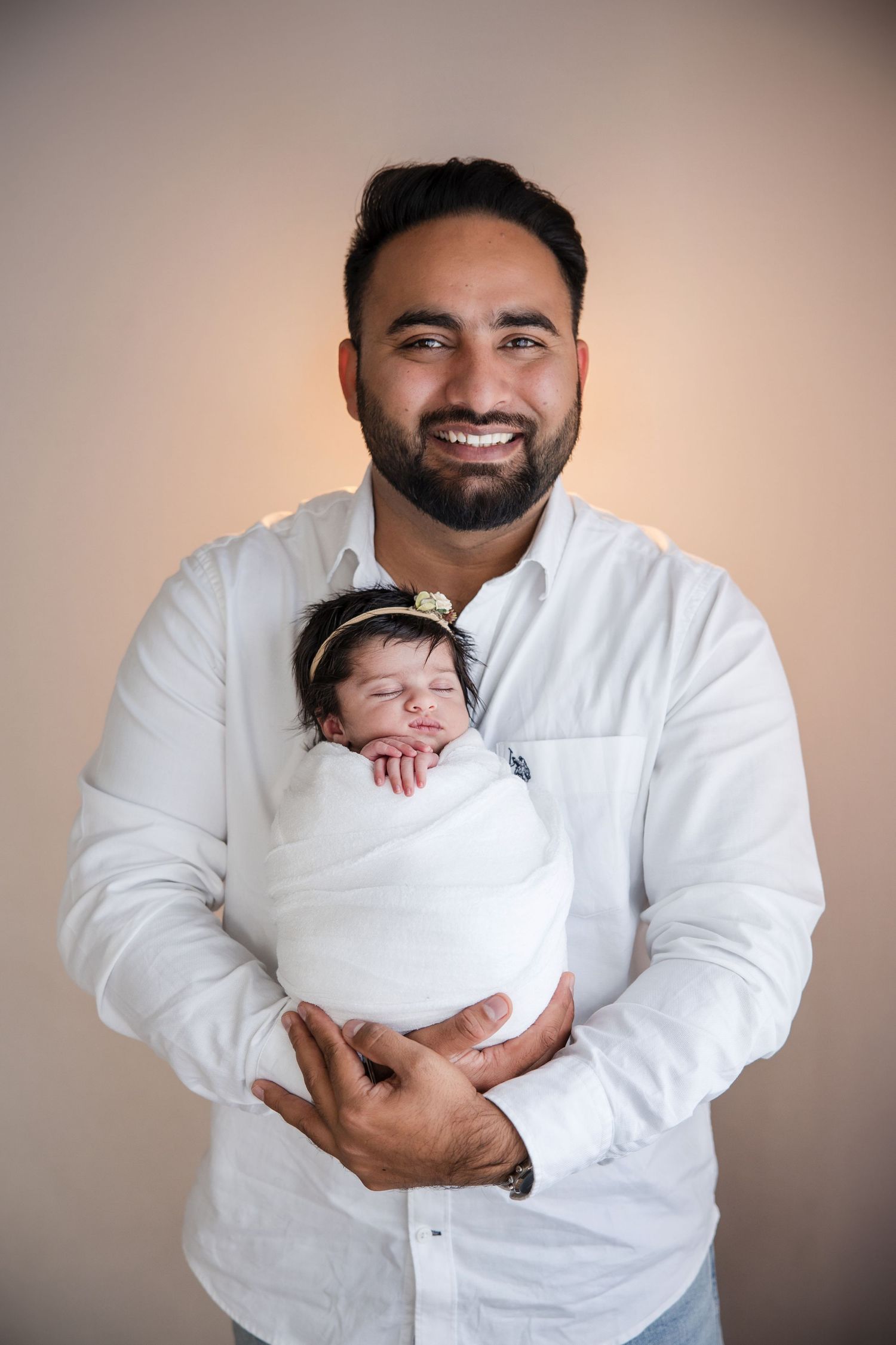 Smiling bearded man in white shirt proudly holds swaddled newborn baby with headband against neutral background.