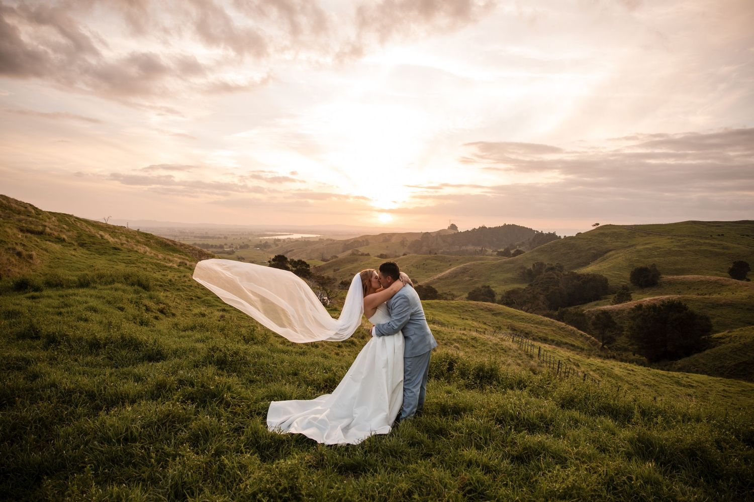 Bride and groom kissing at sunset on green hills, wedding veil flowing in the wind.