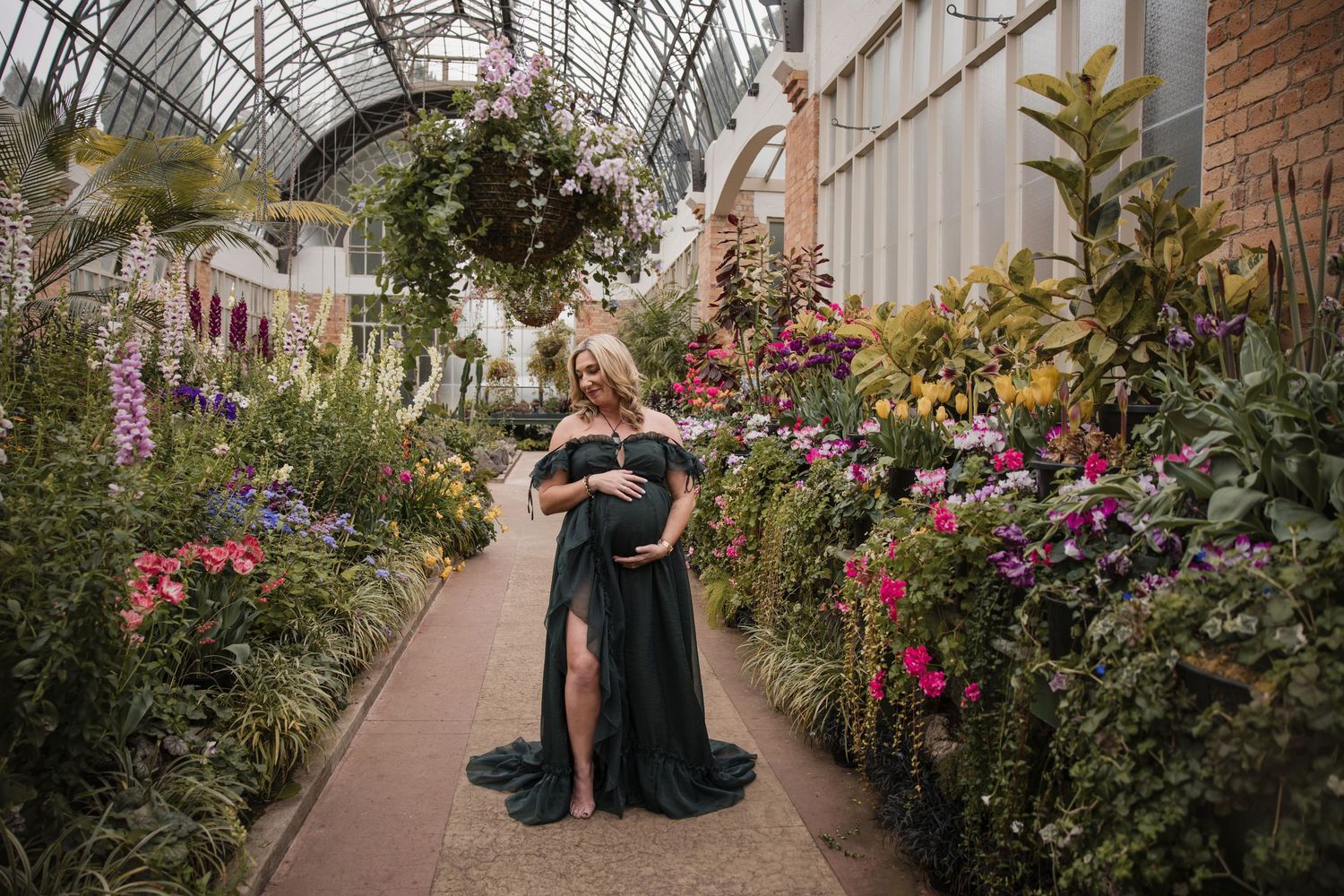 Pregnant woman in elegant dark green gown poses in lush greenhouse surrounded by colorful blooming flowers.