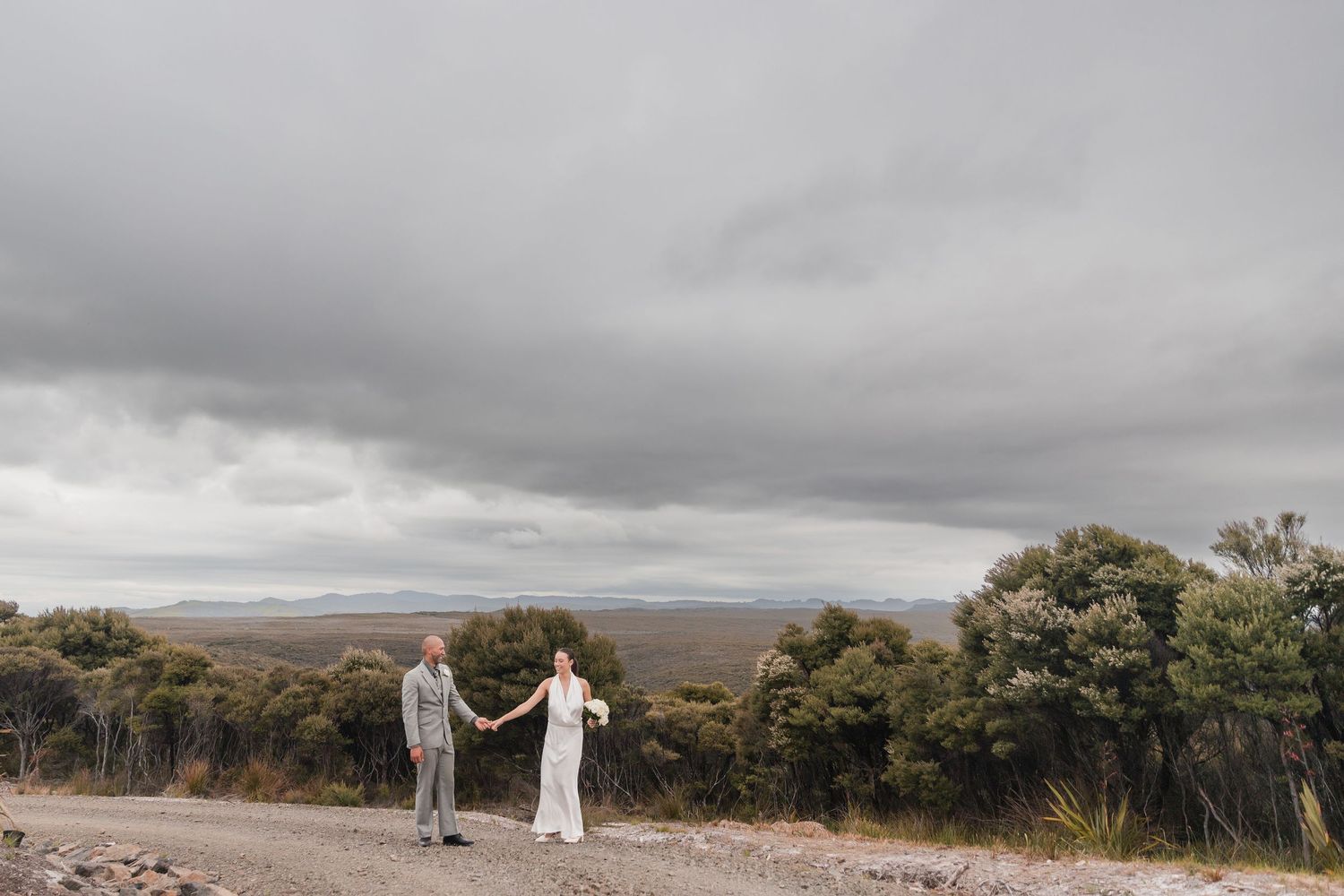 A couple stands holding hands on a scenic mountain overlook under dramatic cloudy skies.