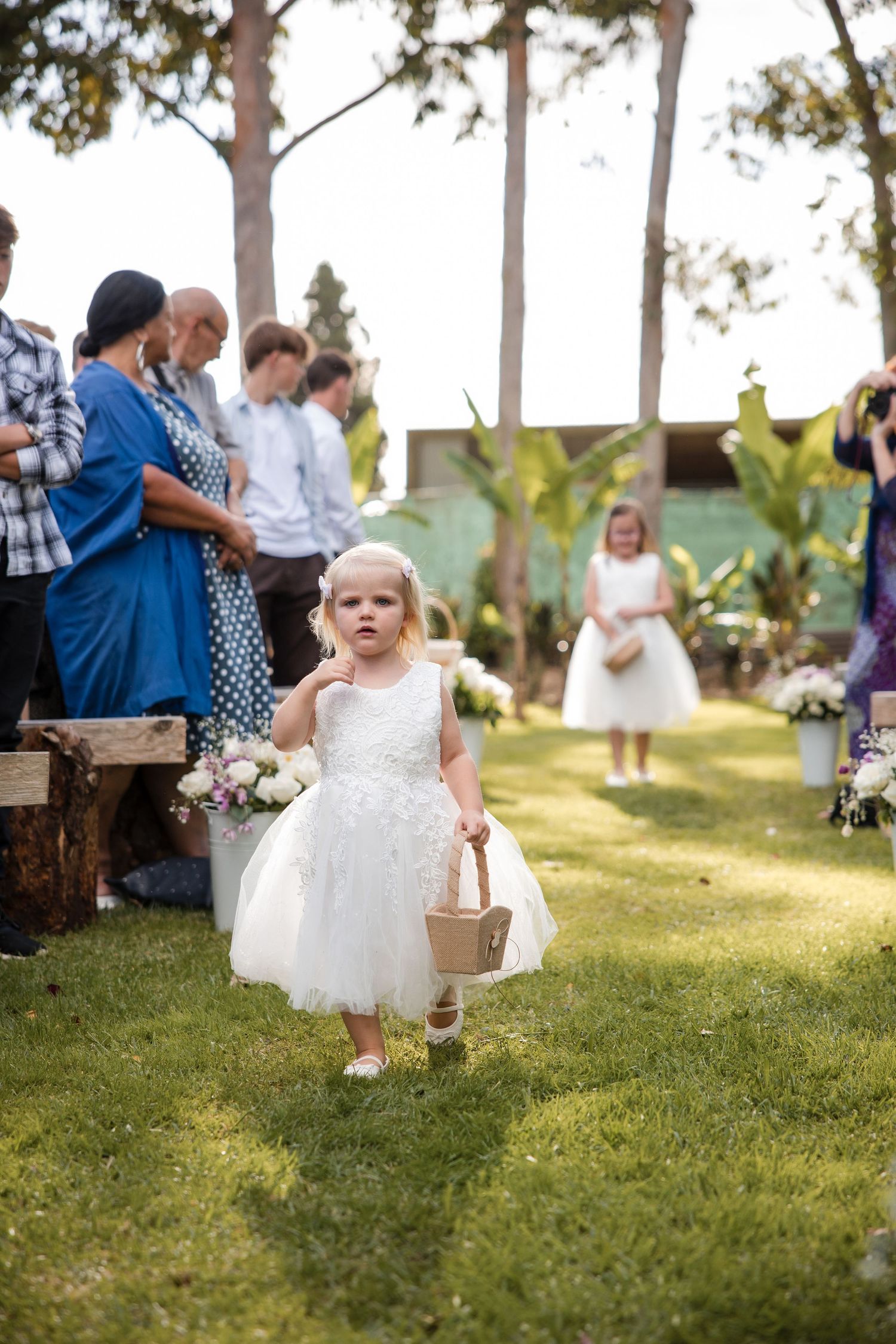 A flower girl in a white dress carries a basket down the grassy aisle during an outdoor wedding ceremony.