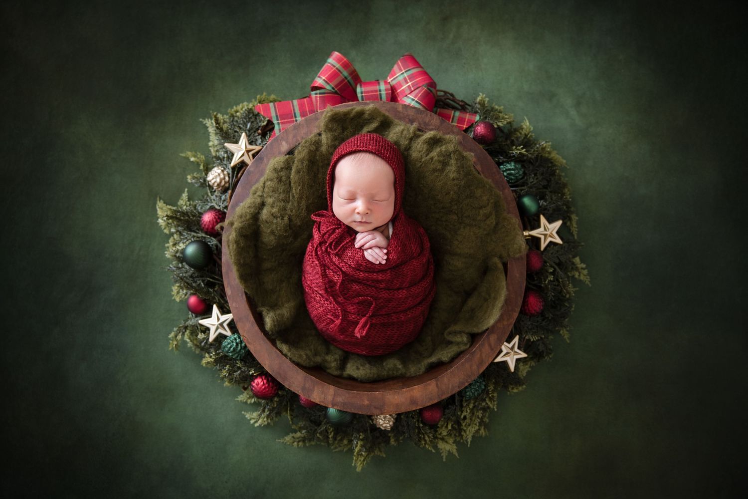 Newborn baby swaddled in red wrap sleeping in a wooden bowl with Christmas wreath decoration and festive ornaments.