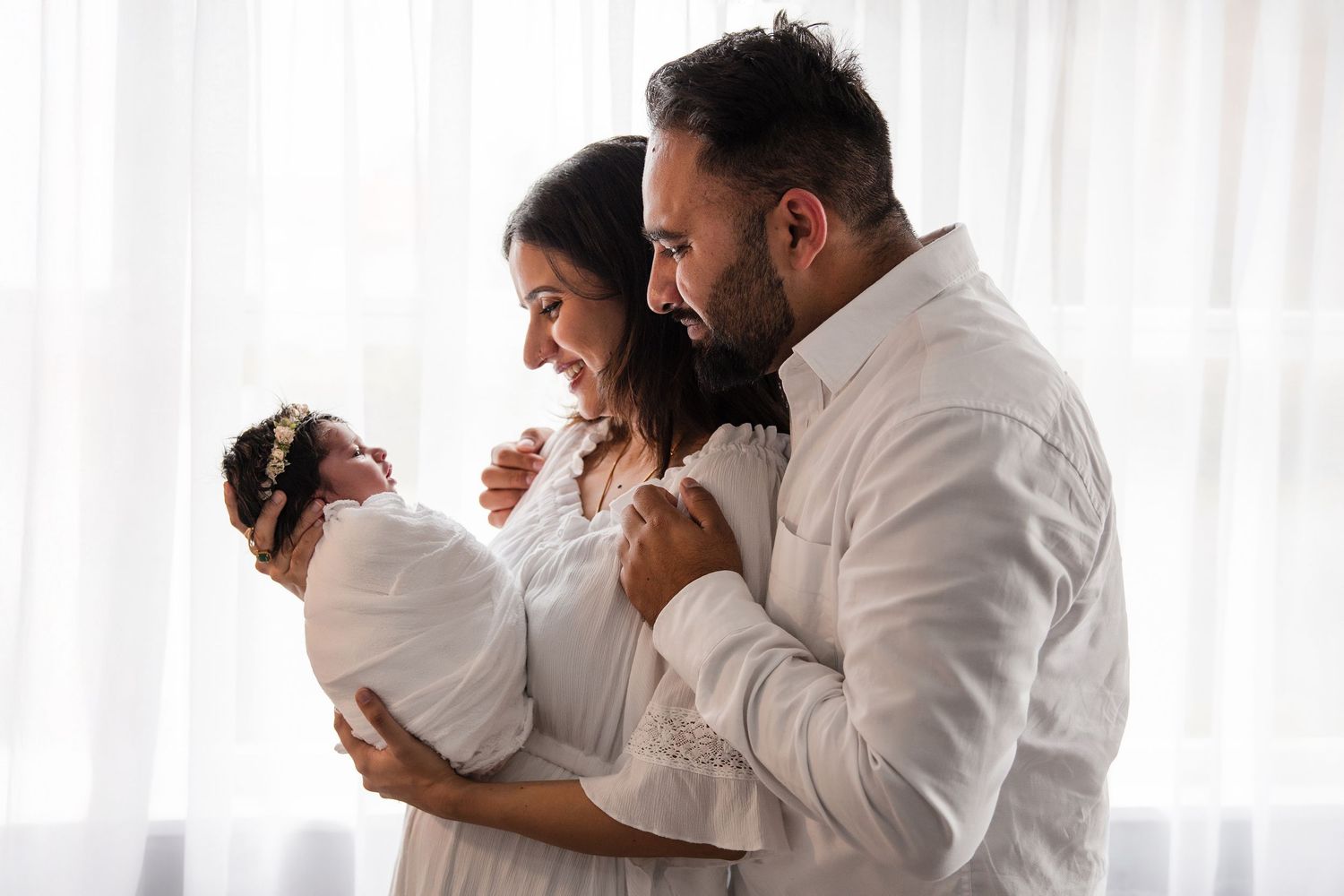 A couple in white clothing admires their newborn baby wrapped in white, standing by a bright window.
