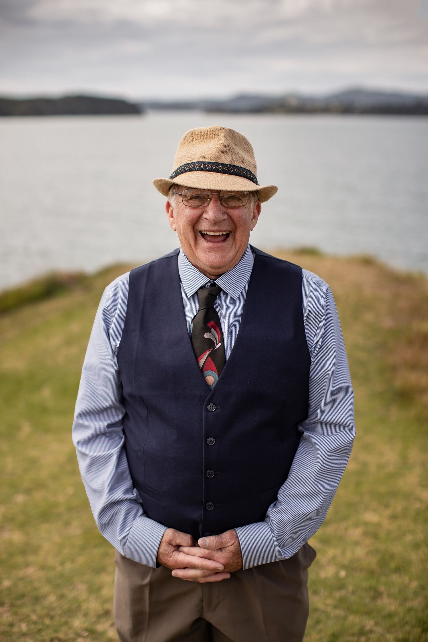 Joyful person in vest and fedora laughing while standing on coastal cliffside with water and hills in background.