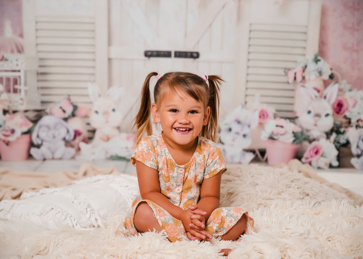 Child in peach floral dress smiling and playing on a soft white rug in front of white shutters.