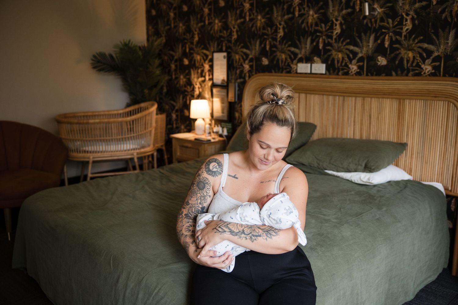 Tattooed mother breastfeeding newborn baby while sitting on bed in stylish hotel room with tropical wallpaper.