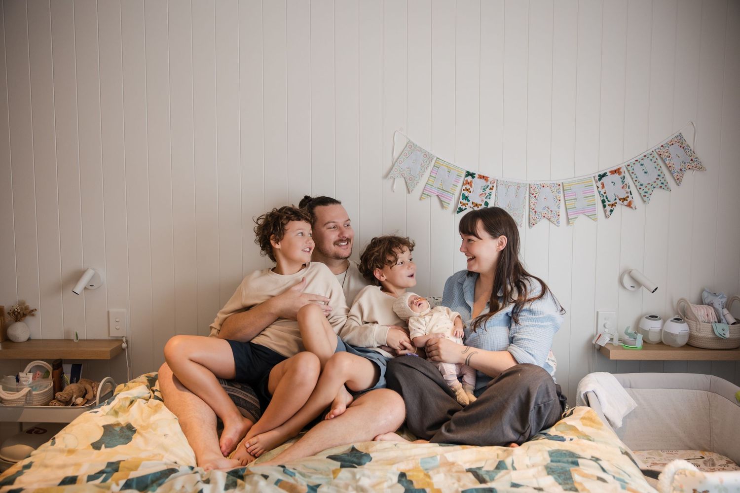 Happy family of four sitting on a bed, laughing together with birthday bunting decorations in a cozy bedroom.