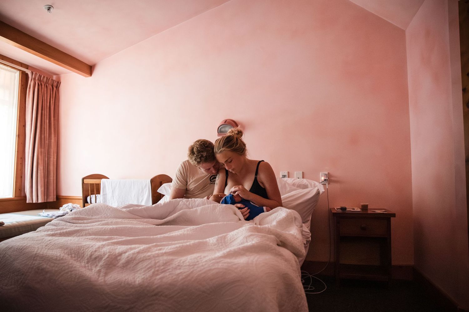 Woman breastfeeding her newborn baby while sitting on a bed in a pink bedroom.