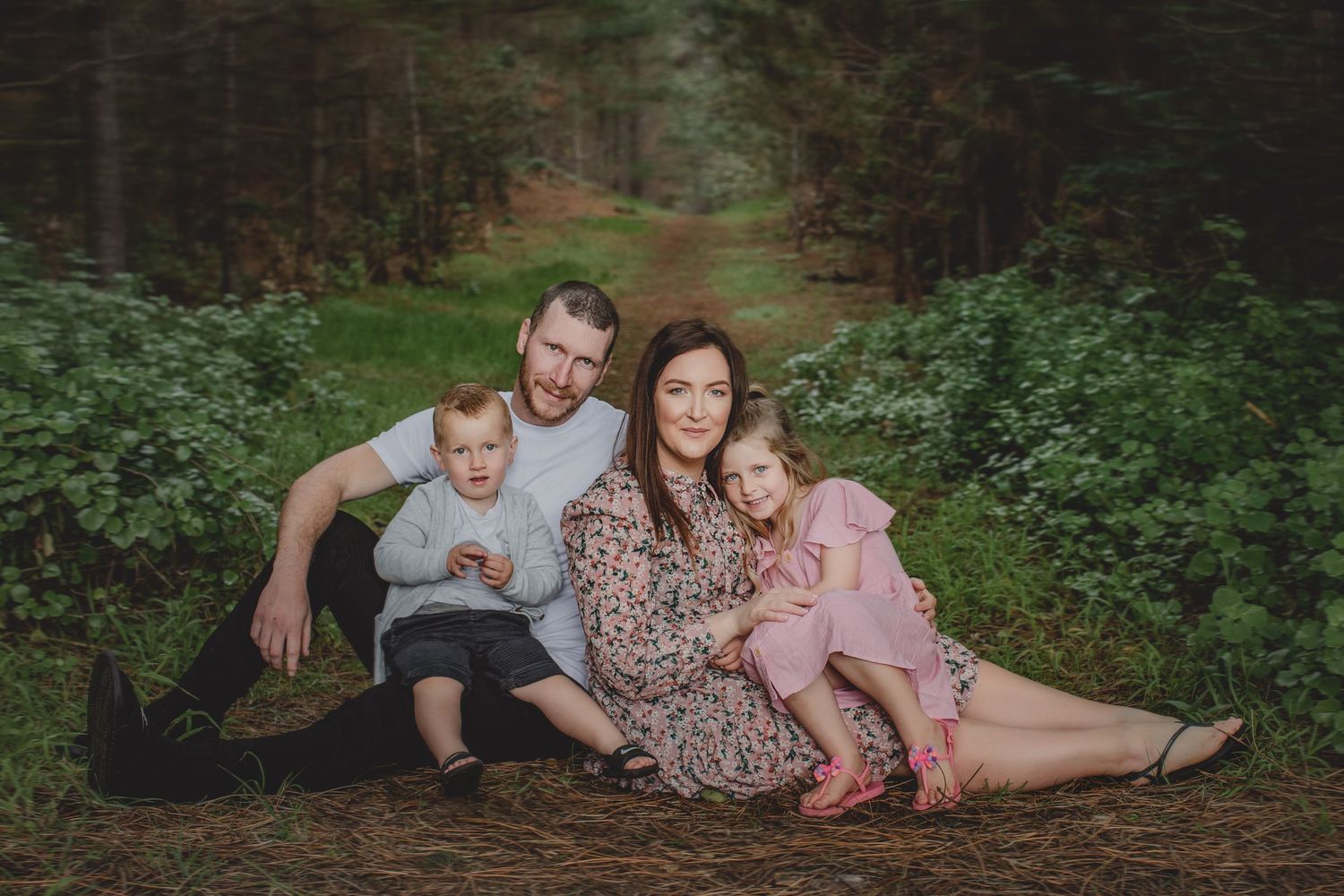 Family relaxes together on forest trail surrounded by green foliage.
