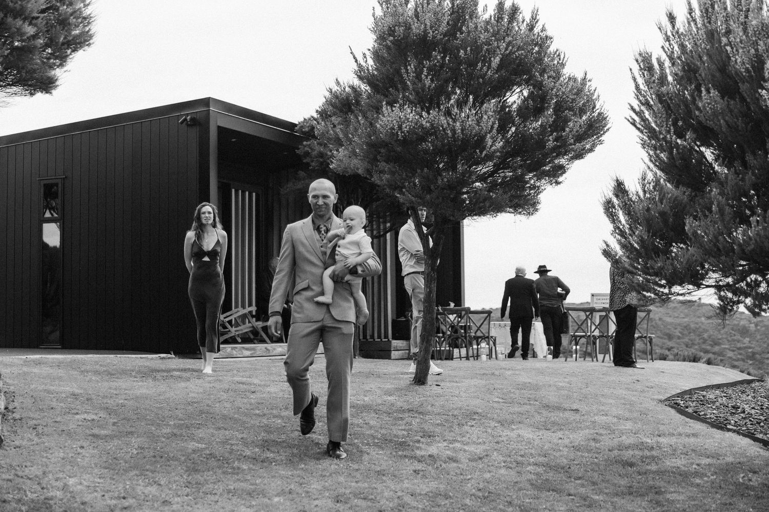 A couple walks along rolling grassy hills at an outdoor venue with pine trees in the background.
