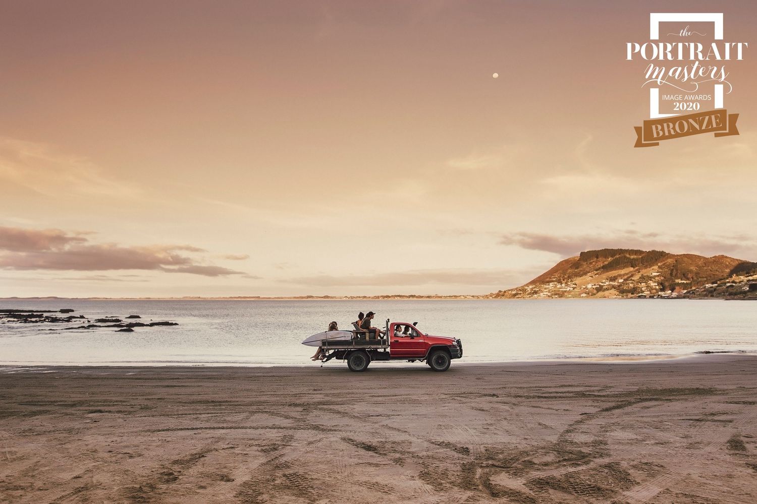 A 4x4 vehicle traverses across sandy beach terrain at sunset with rolling waves in the background.