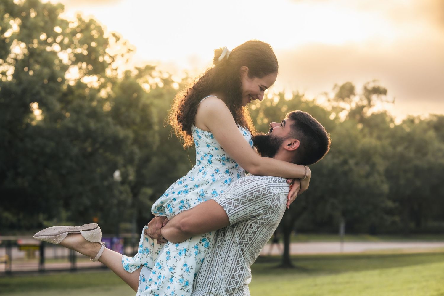 A man lifts a woman in a floral dress outdoors at sunset, sharing a romantic moment in a lush green park.