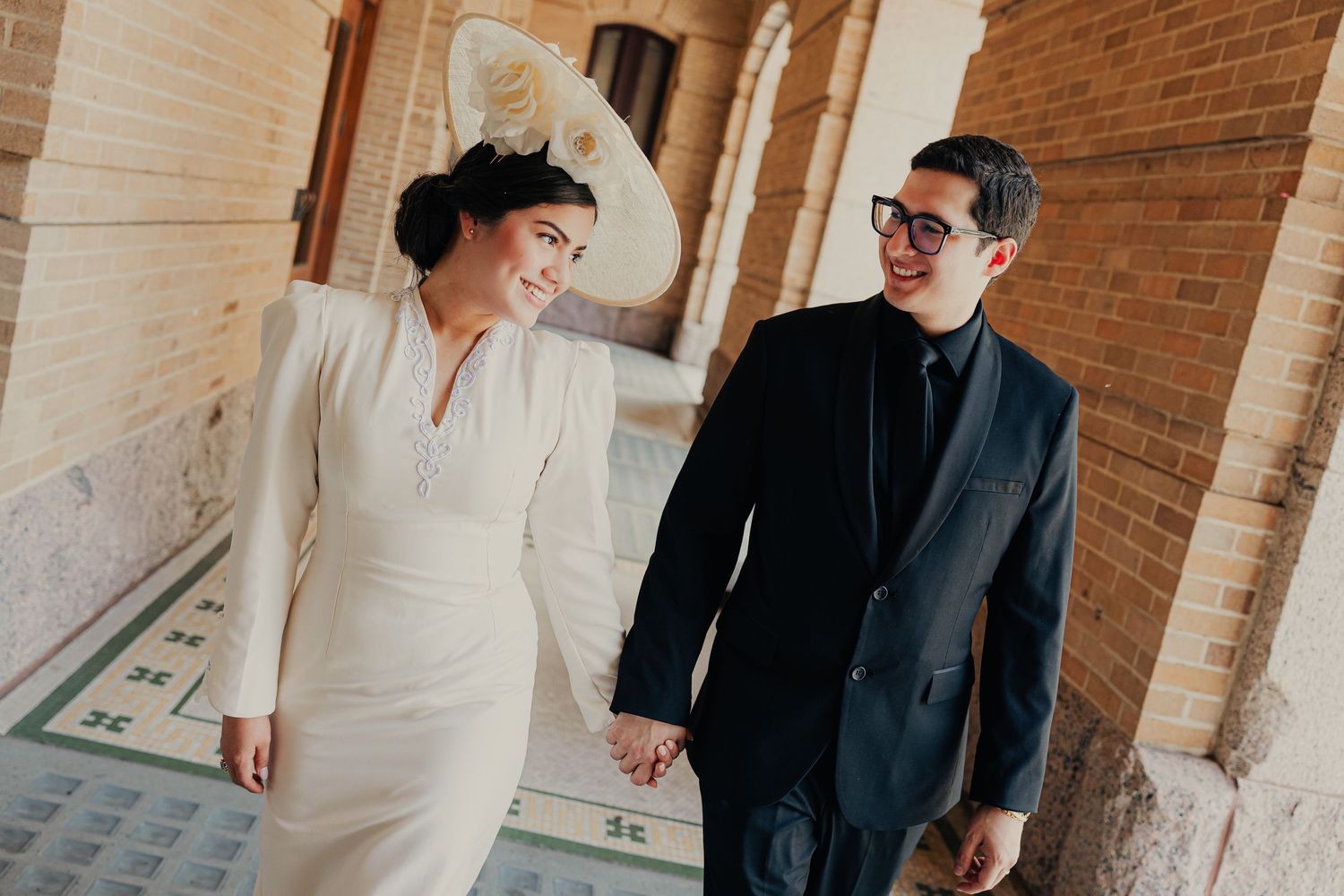 Couple holding hands walking through brick archway, woman in white dress and hat, man in black suit.