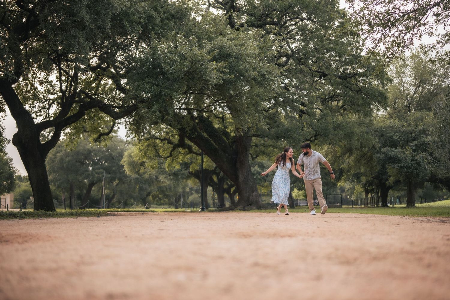 Couple walking hand in hand through a lush green park with large oak trees on a warm sunny day.