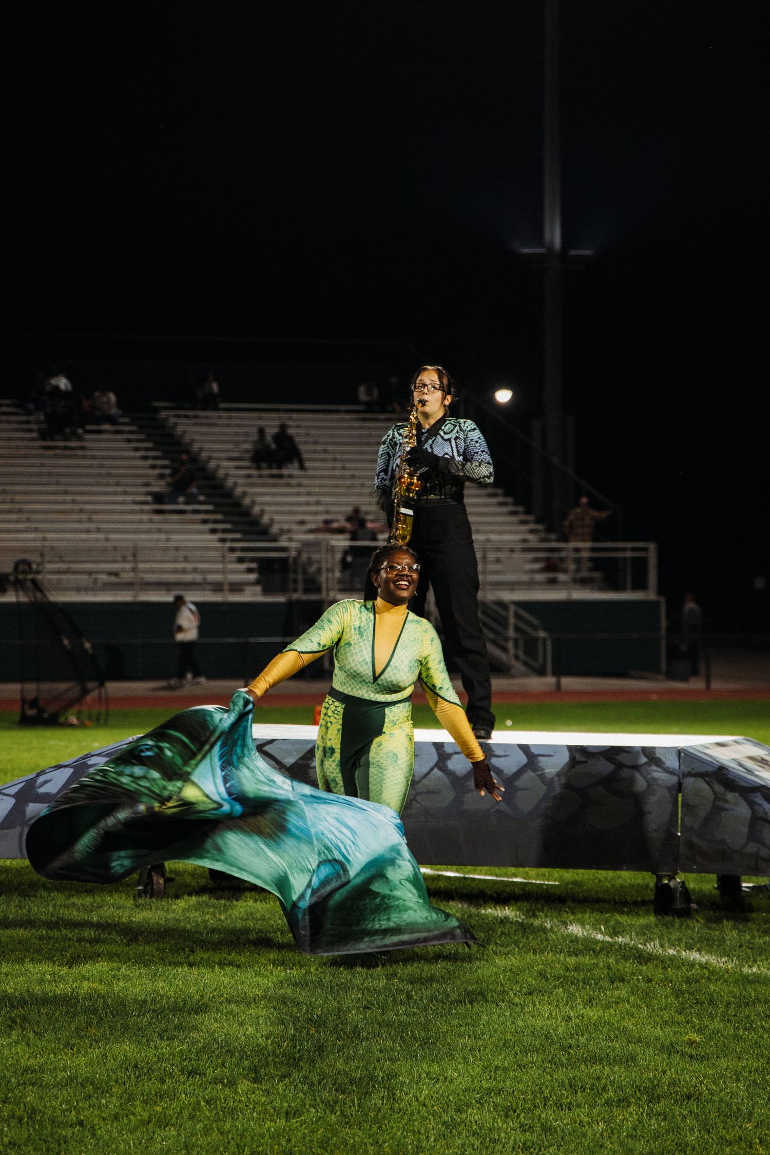 Marching band performers in colorful uniforms lay on the grass field during a nighttime performance.