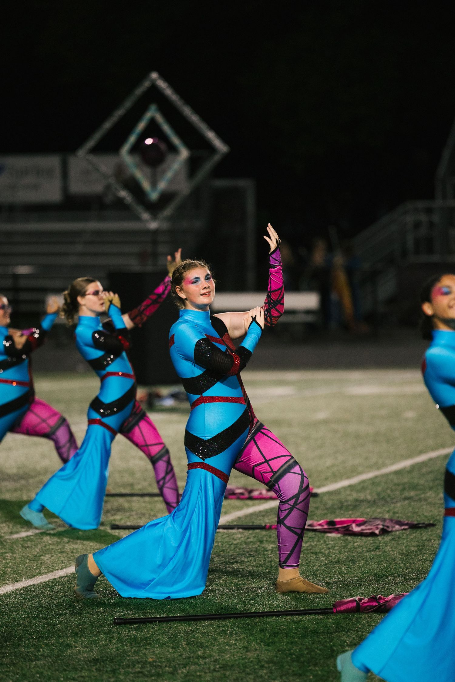 Color guard performers in blue and pink uniforms execute precise routines on a football field during a nighttime competition.