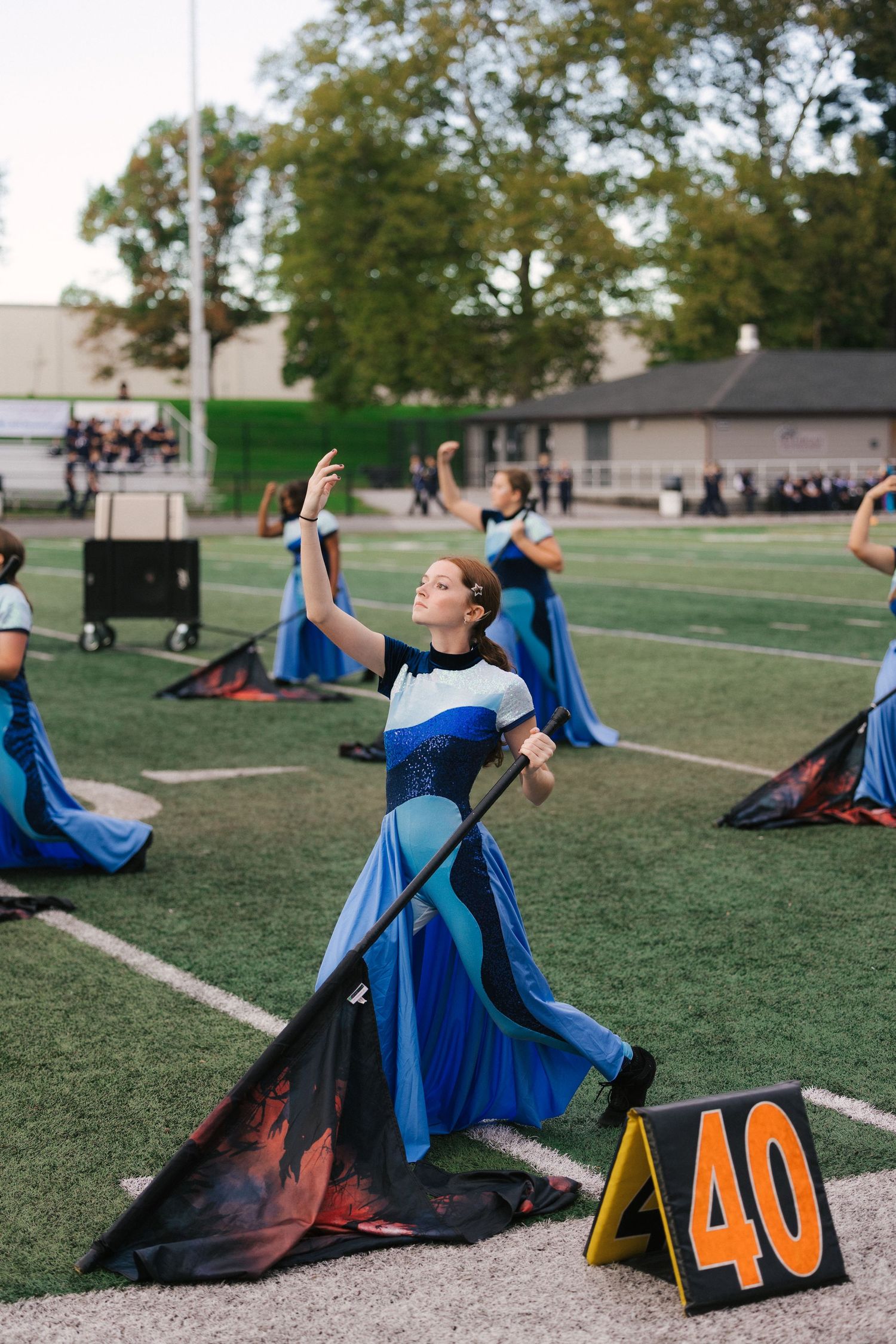 Color guard performer in blue costume spinning a flag on a football field at a marching band competition.