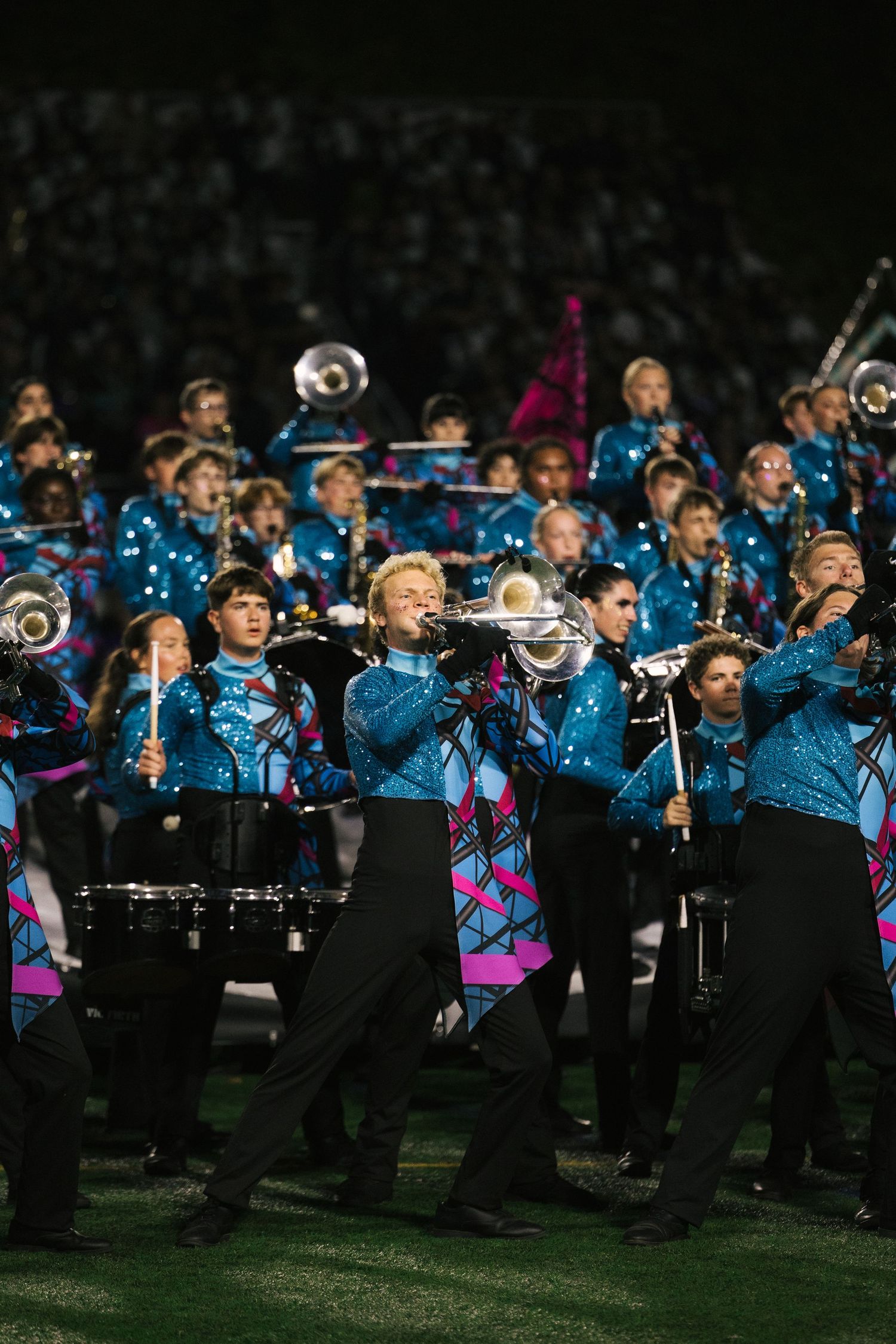 Marching band performing in blue sequined uniforms playing brass instruments on a football field at night.