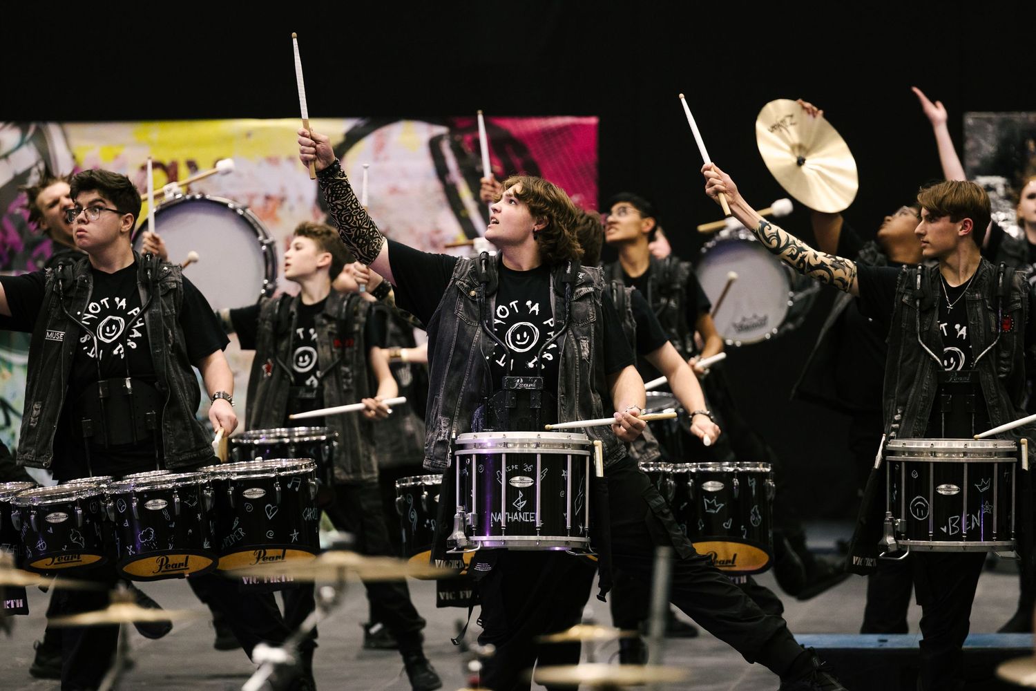 Young drummers in black uniforms perform energetically on snare drums and cymbals during a competitive indoor percussion event.