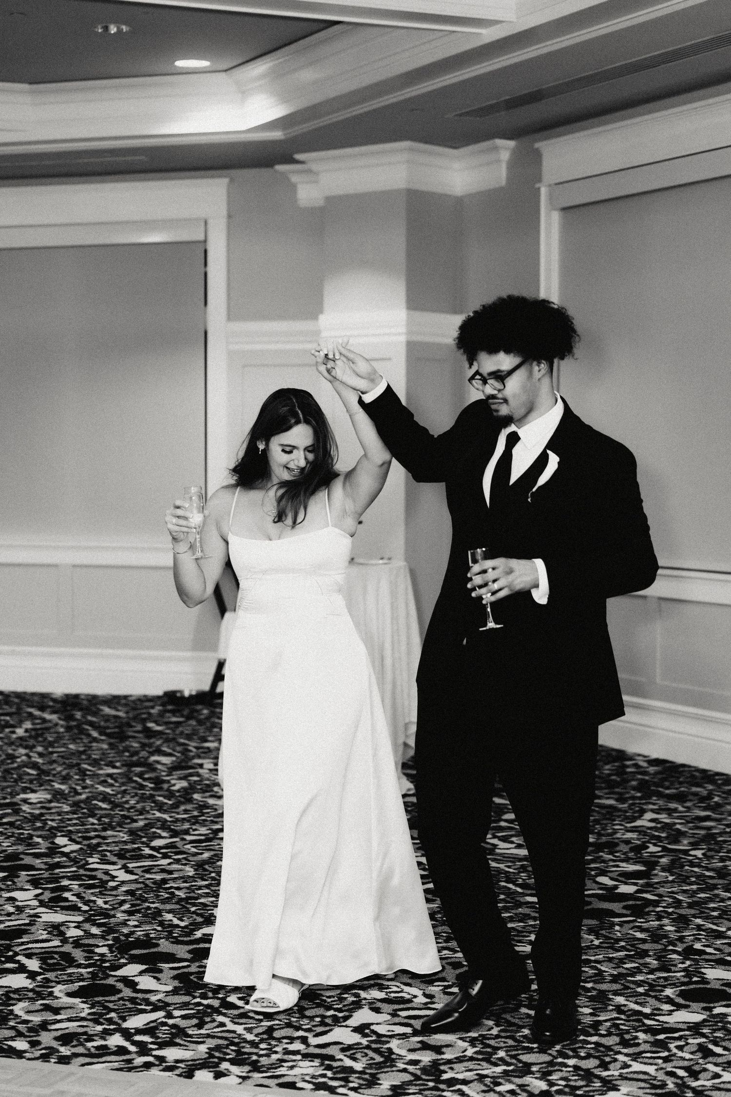 A couple dancing together at a wedding reception, with the bride in white and groom in black suit holding champagne glasses.