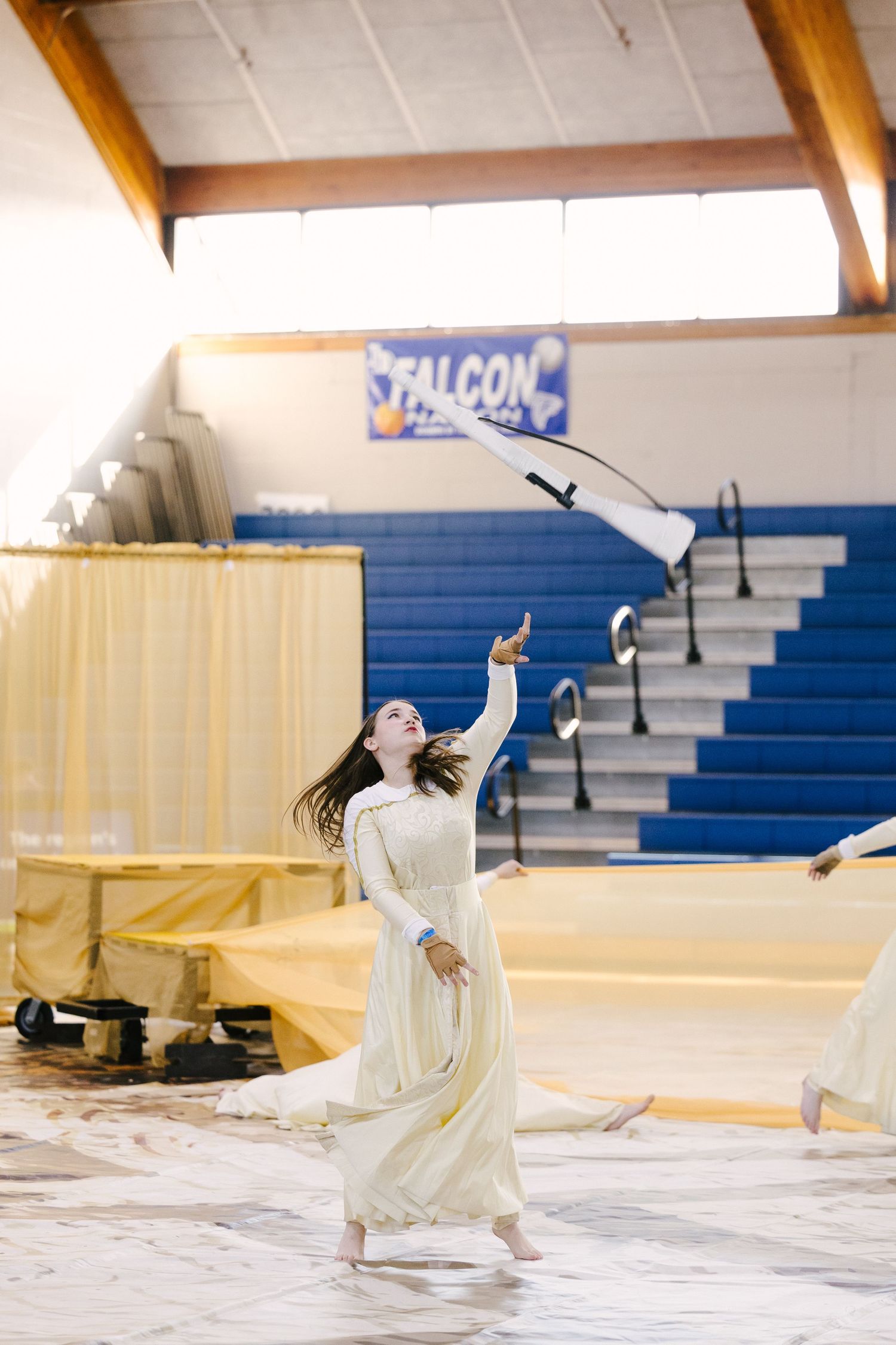 Barefoot winterguard performer in a flowing cream dress tosses a white flag in a school gym with blue bleachers and a Falcon banner.