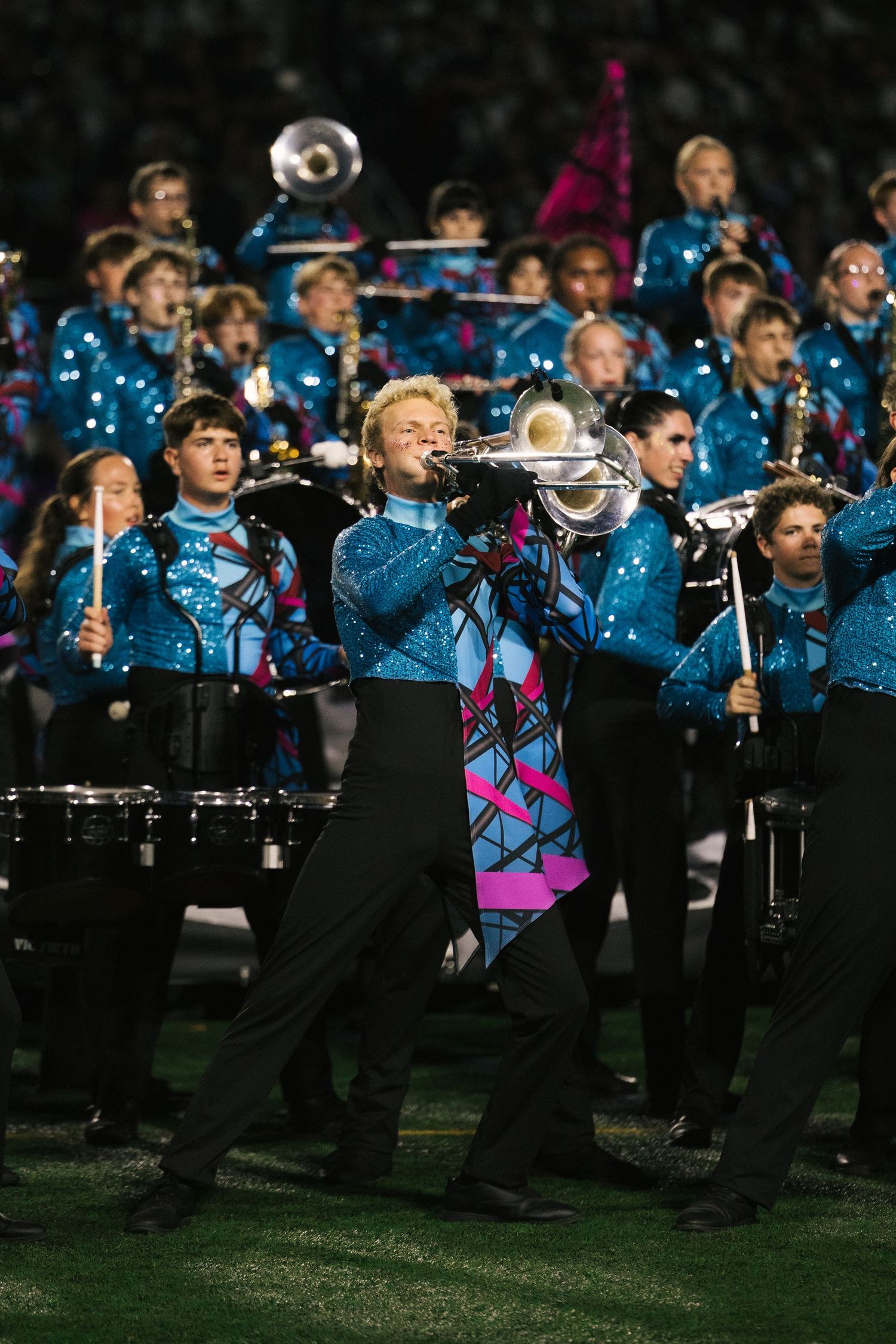 Marching band performers in blue sequin uniforms playing brass instruments during a nighttime field show performance.