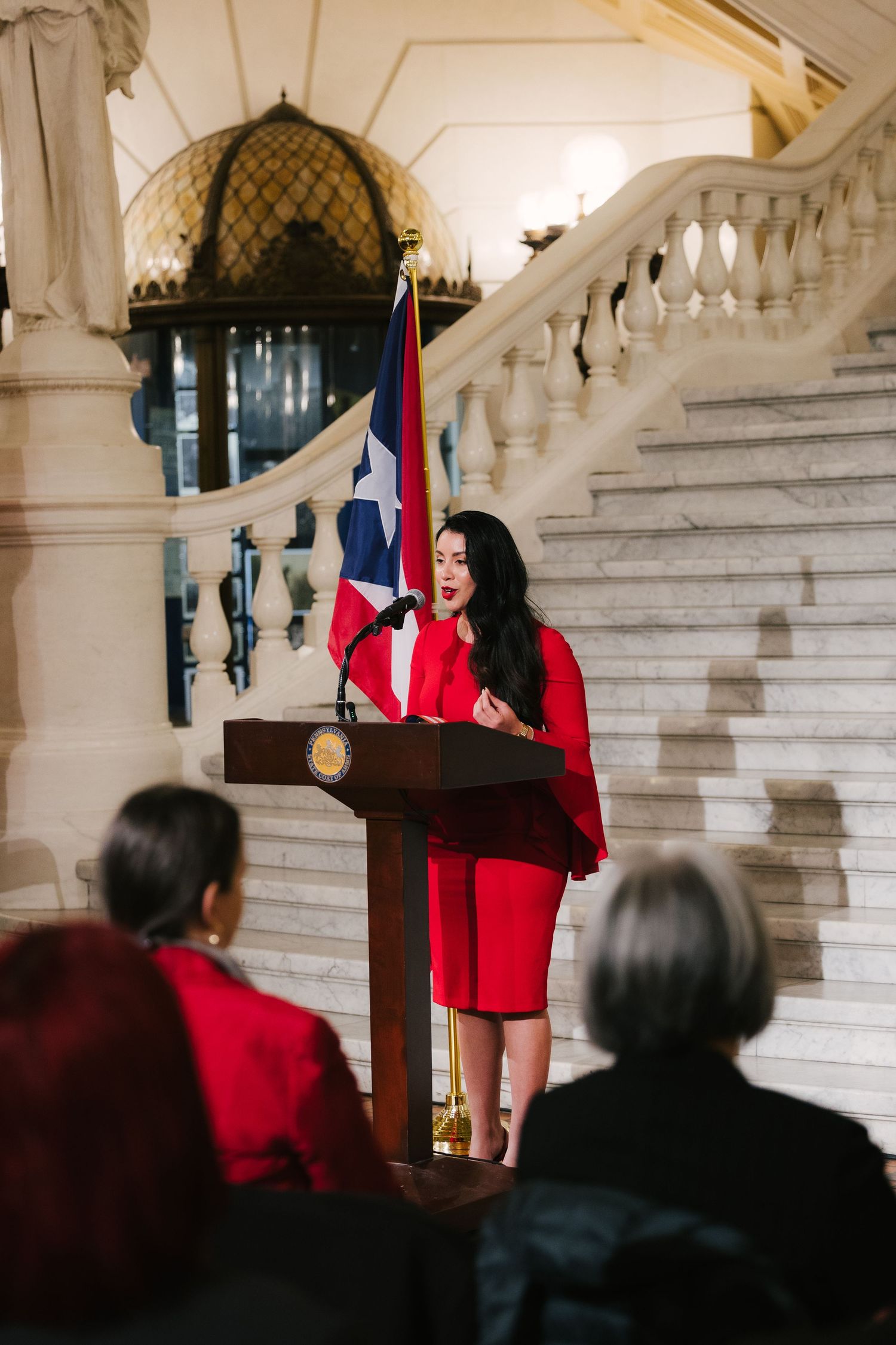 A speaker in a red outfit stands at a podium on a grand white marble staircase during a formal event.