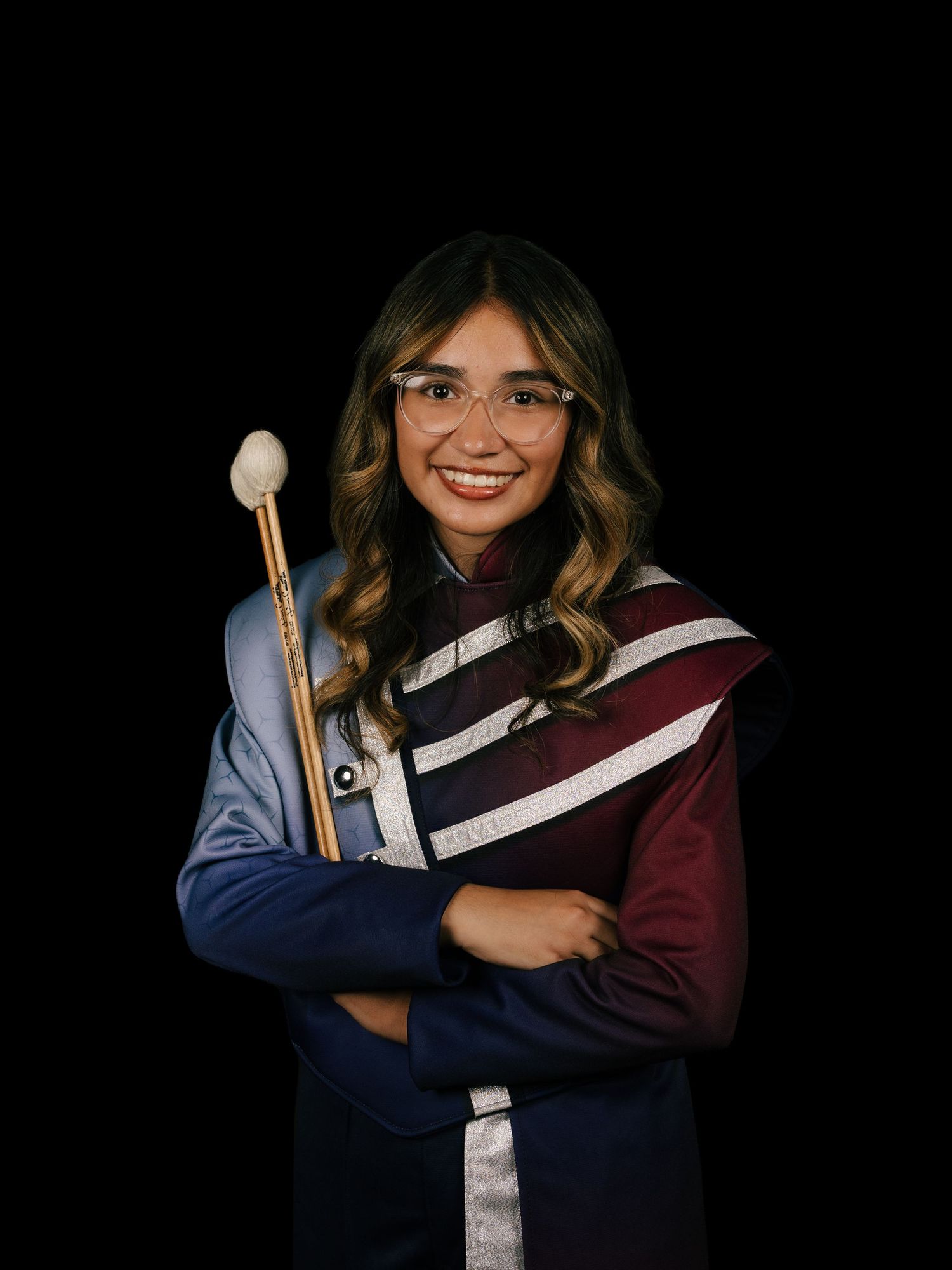 Student in graduation attire wearing glasses holds a band conductor's baton against a dark background.