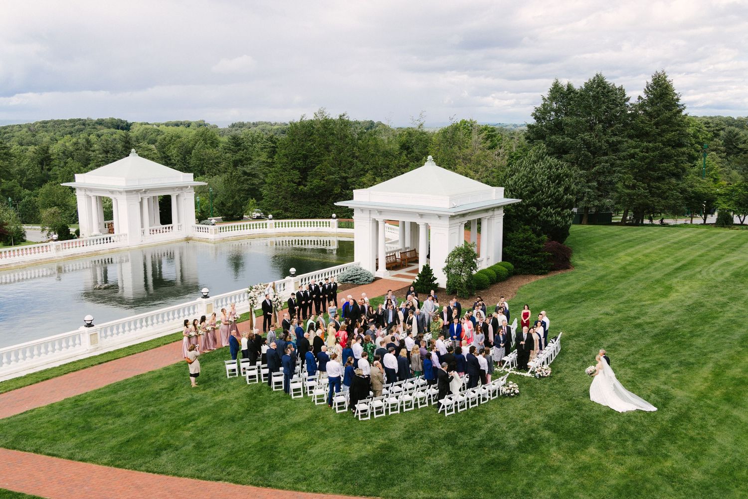 A panoramic view of an elegant outdoor wedding ceremony with white gazebos and guests seated on a manicured green lawn.