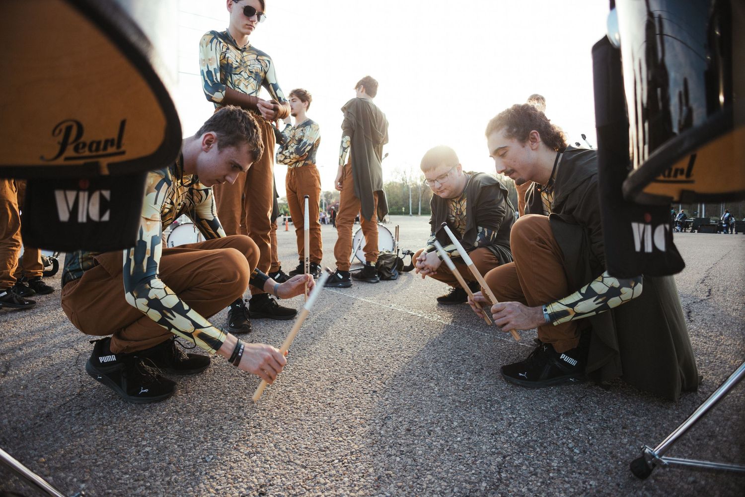 Drumline members in matching uniforms crouch on pavement tuning Pearl drums at sunset while teammates stand in the background.