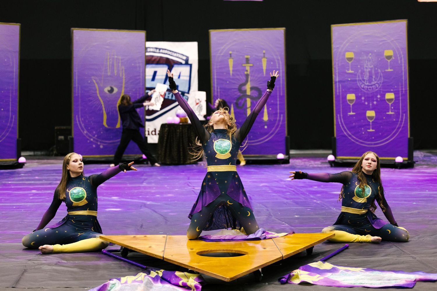 Color guard performers in purple and gold costumes execute a dramatic floor routine on a competition stage.