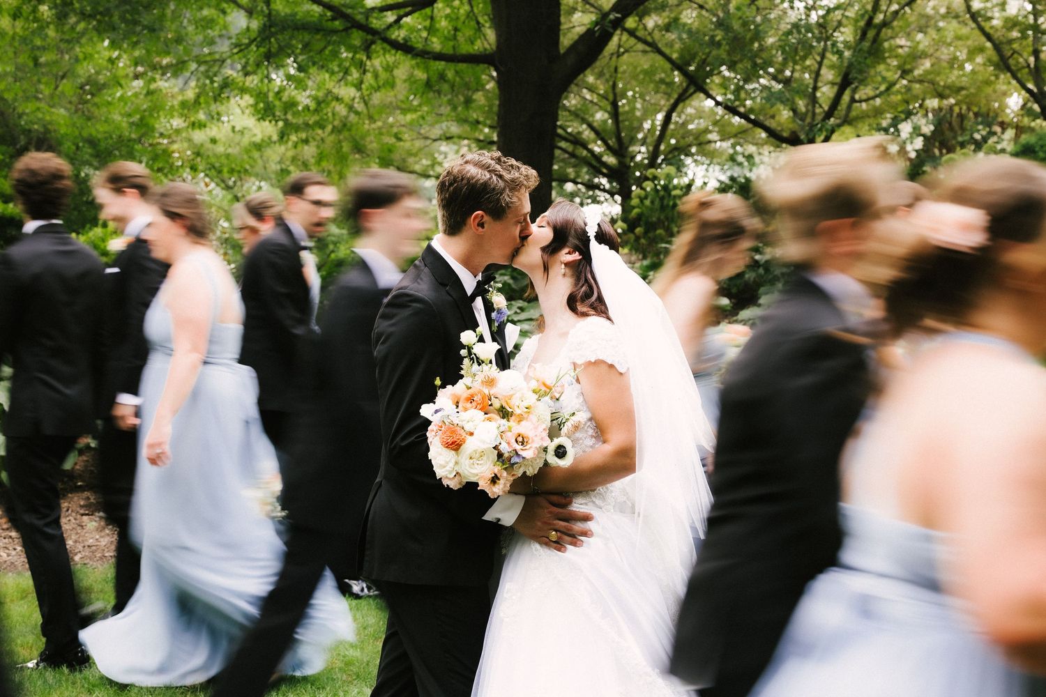 Motion-blurred wedding party walking through a lush green garden setting.