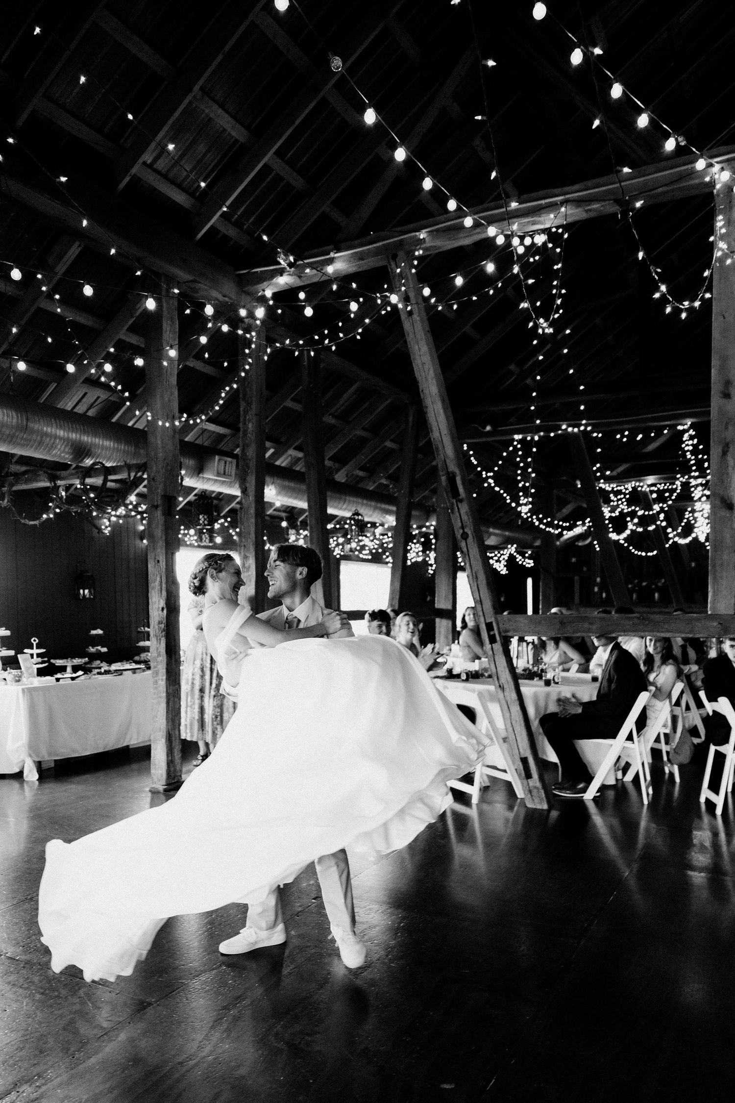 A wedding dance floor in a rustic barn venue illuminated by string lights showing flowing white dress movement.