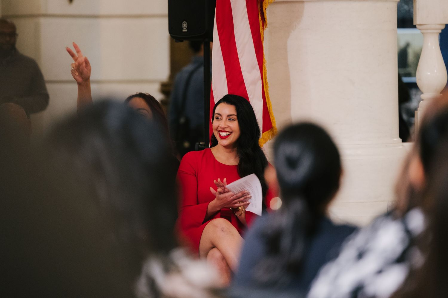 A person in a red dress gestures while speaking to a crowd at a podium with an American flag in the background.