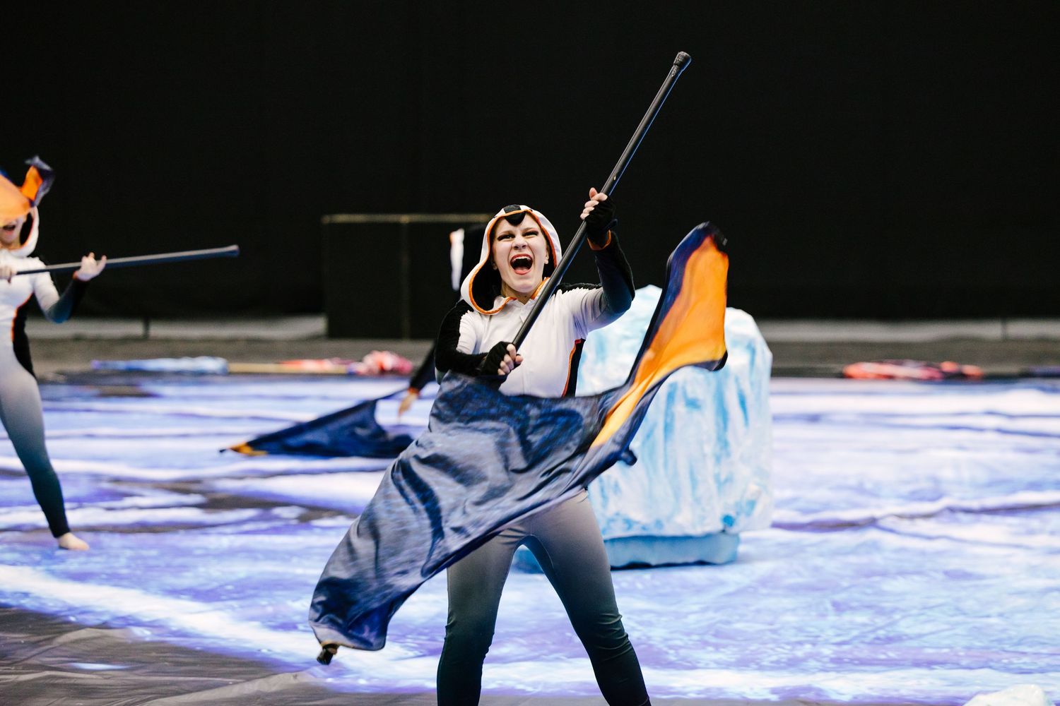 Indoor color guard members practice with flags and equipment on a blue performance floor.