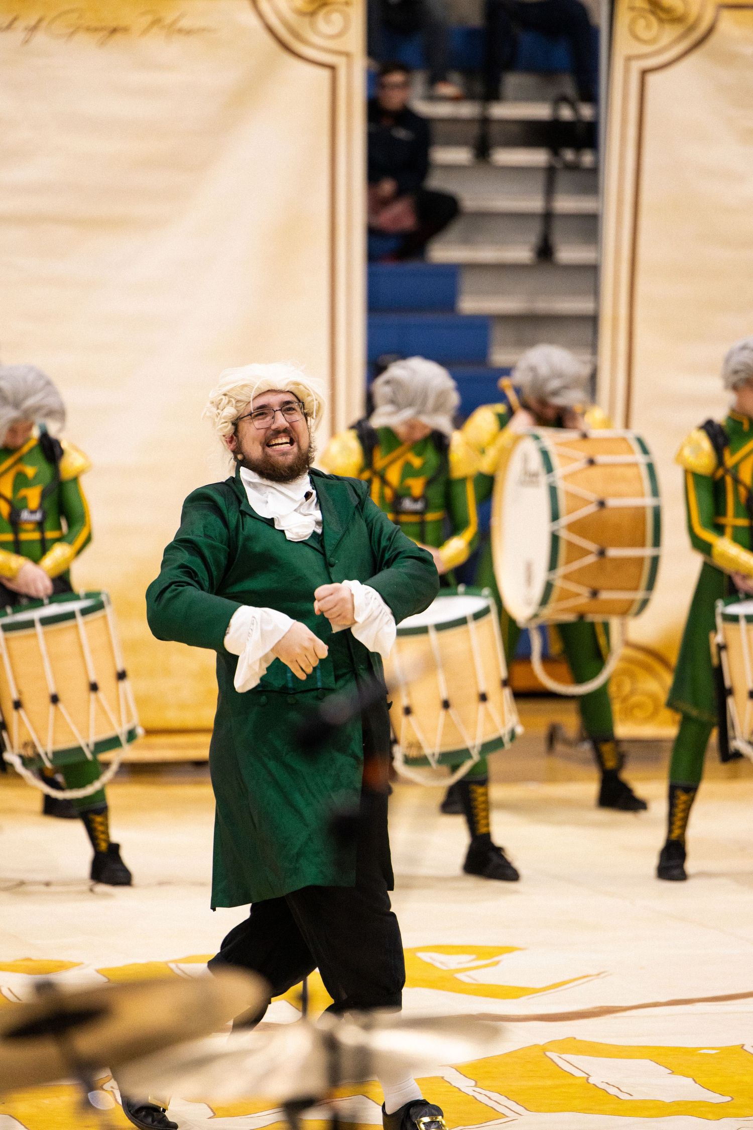 Performer in green period costume dances energetically across a yellow stage floor.
